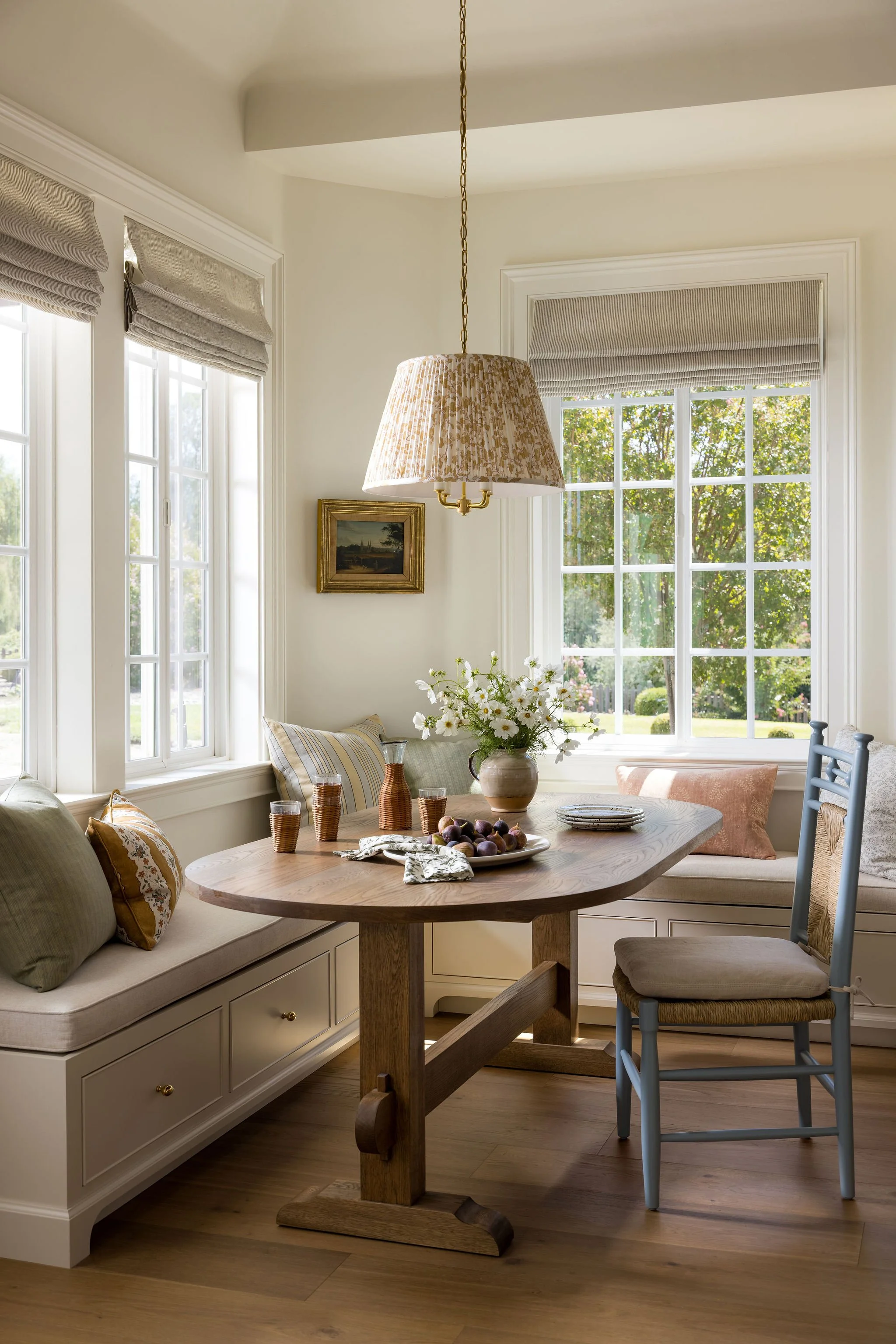 A cozy dining nook with large windows, a wooden table with decorative items, a built-in window seat with pillows, and a hanging pendant lamp with a patterned shade.