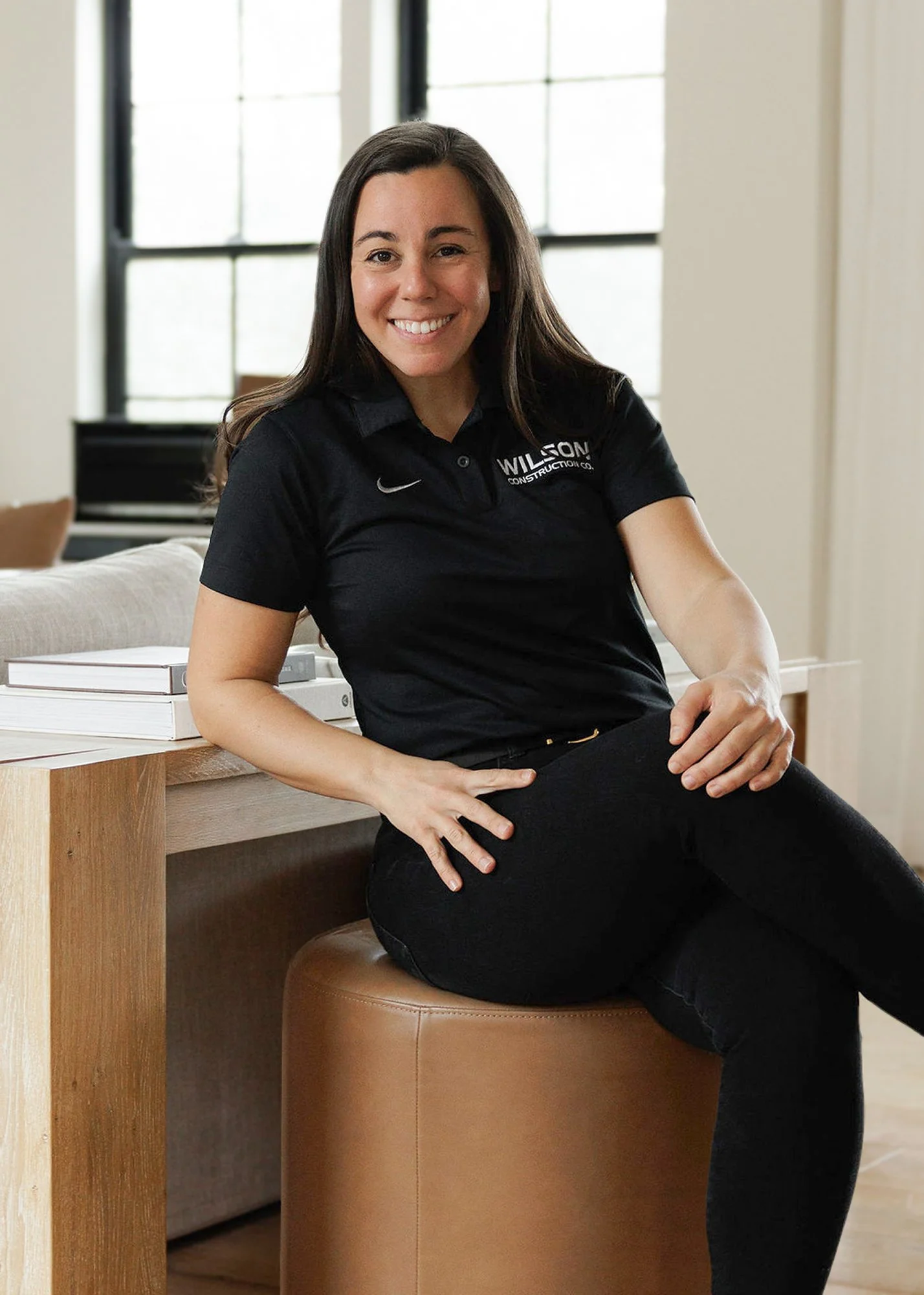 A woman with long brown hair, smiling, sitting on a tan ottoman, wearing a black polo shirt with a logo, in a bright room with large windows.