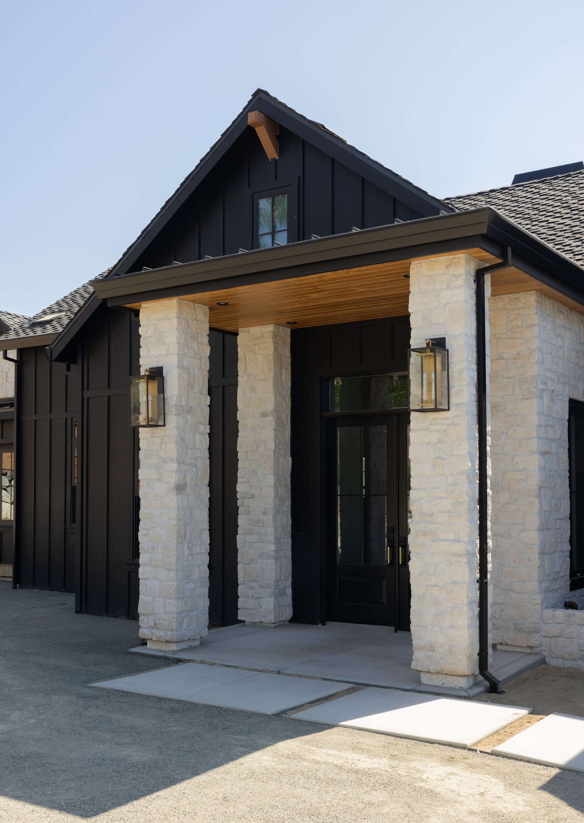 Modern house with black siding, white brick columns, a wooden porch ceiling, and outdoor lighting fixtures.