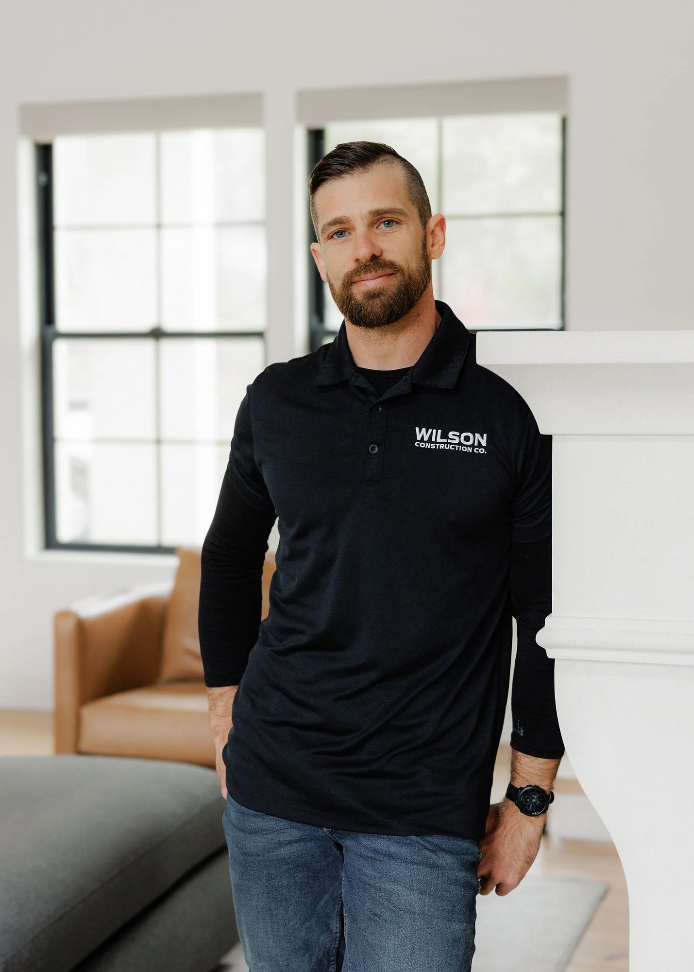 A man standing indoors, wearing a black polo shirt with 'Wilson Construction Co.' embroidered on it. He has a beard, short hair, and a watch on his left wrist. The background features large windows and a brown chair.