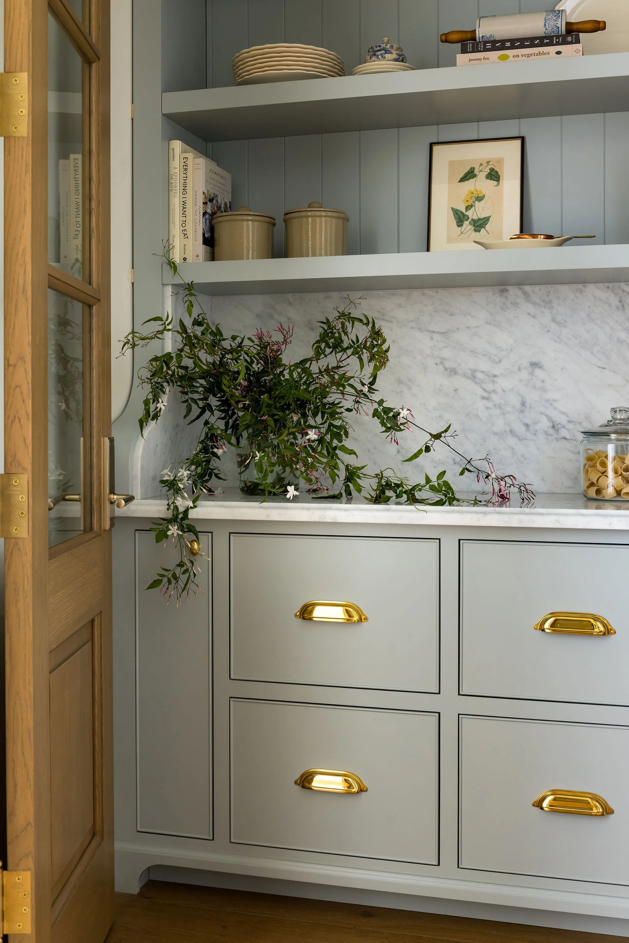 A kitchen shelf with stacked plates, blue and white ceramic jar, rolling pin, and cookbooks. Below the shelf is a light blue cabinet with gold handles, and a marble backsplash with a plant in a glass container.