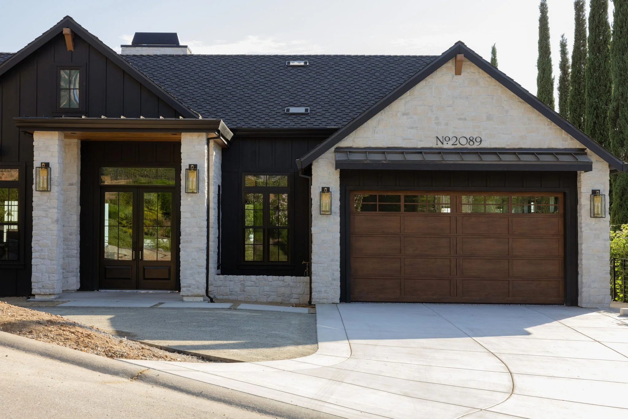 Front view of a modern house with a dark shingled roof, white stone and black siding, and a wooden garage door. Number 2089 displayed on the front wall. Tall cypress trees in the background.