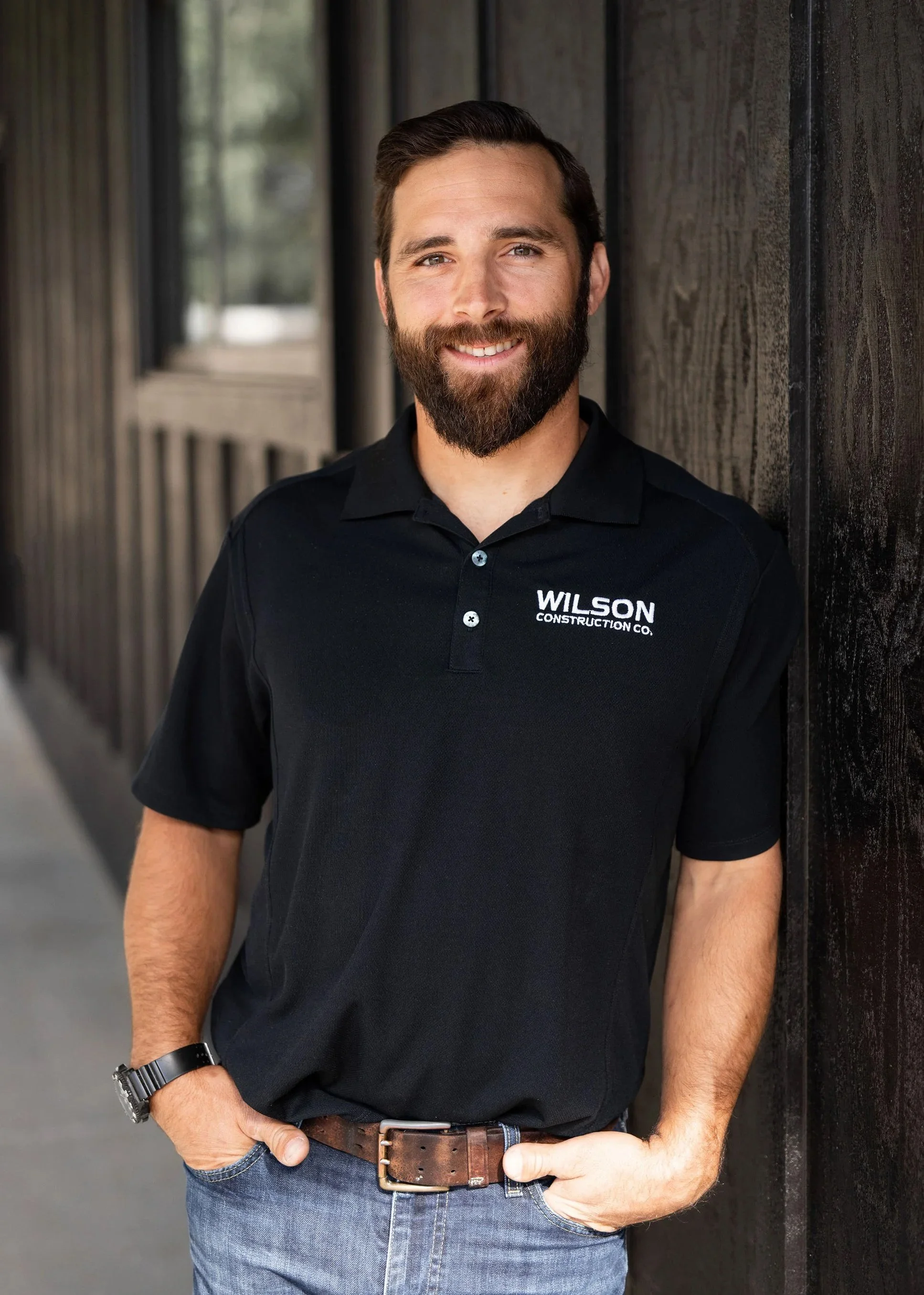Man with a beard and short hair smiling, wearing a black polo shirt with 'Wilson Construction Co.' logo, standing outdoors against a wooden wall.