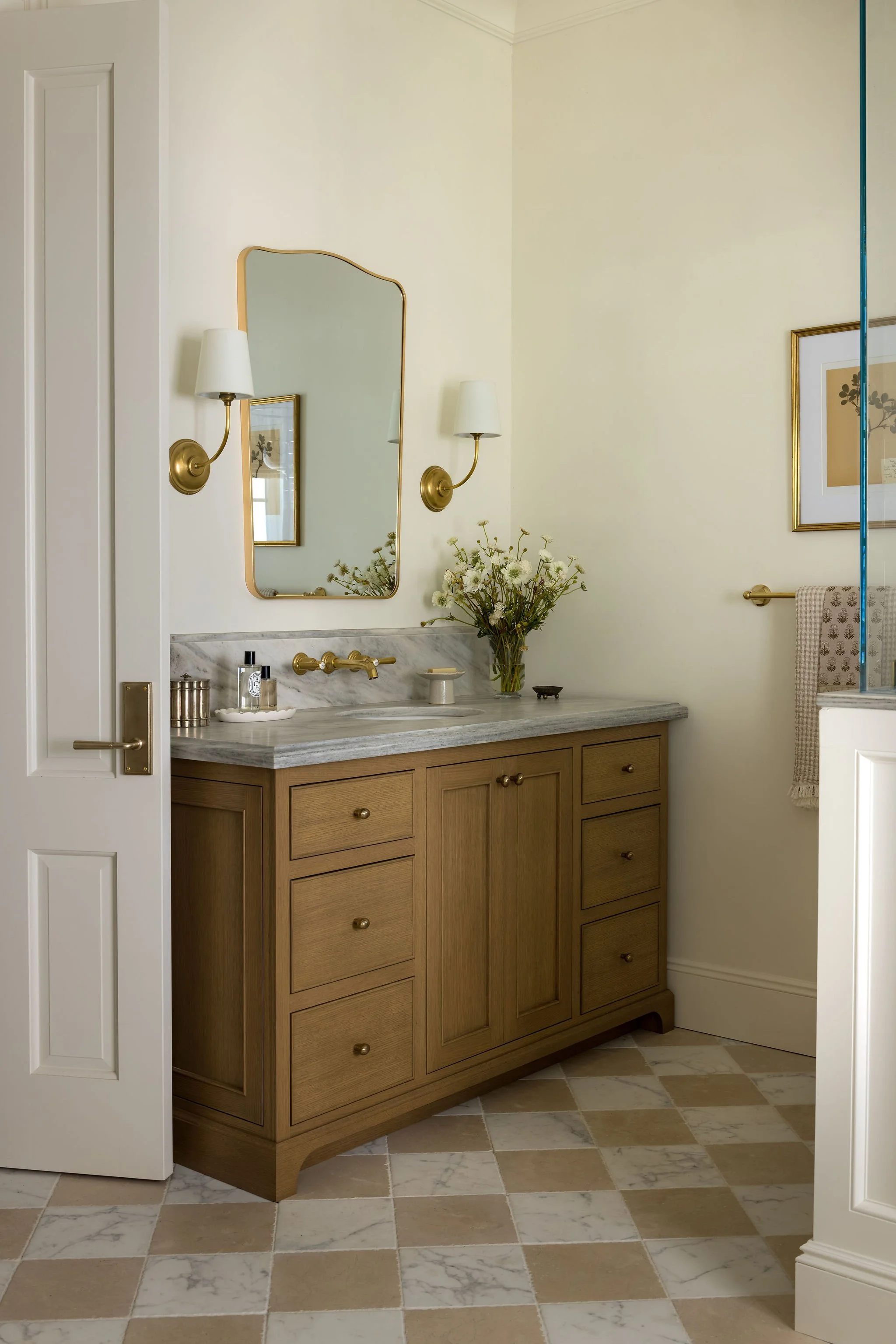 A bathroom vanity area with a wooden cabinet, marble countertop, and a large mirror. Two wall-mounted brass sconces with white shades flank the mirror. There is a framed artwork, a vase with flowers, and some small toiletries on the countertop. The f