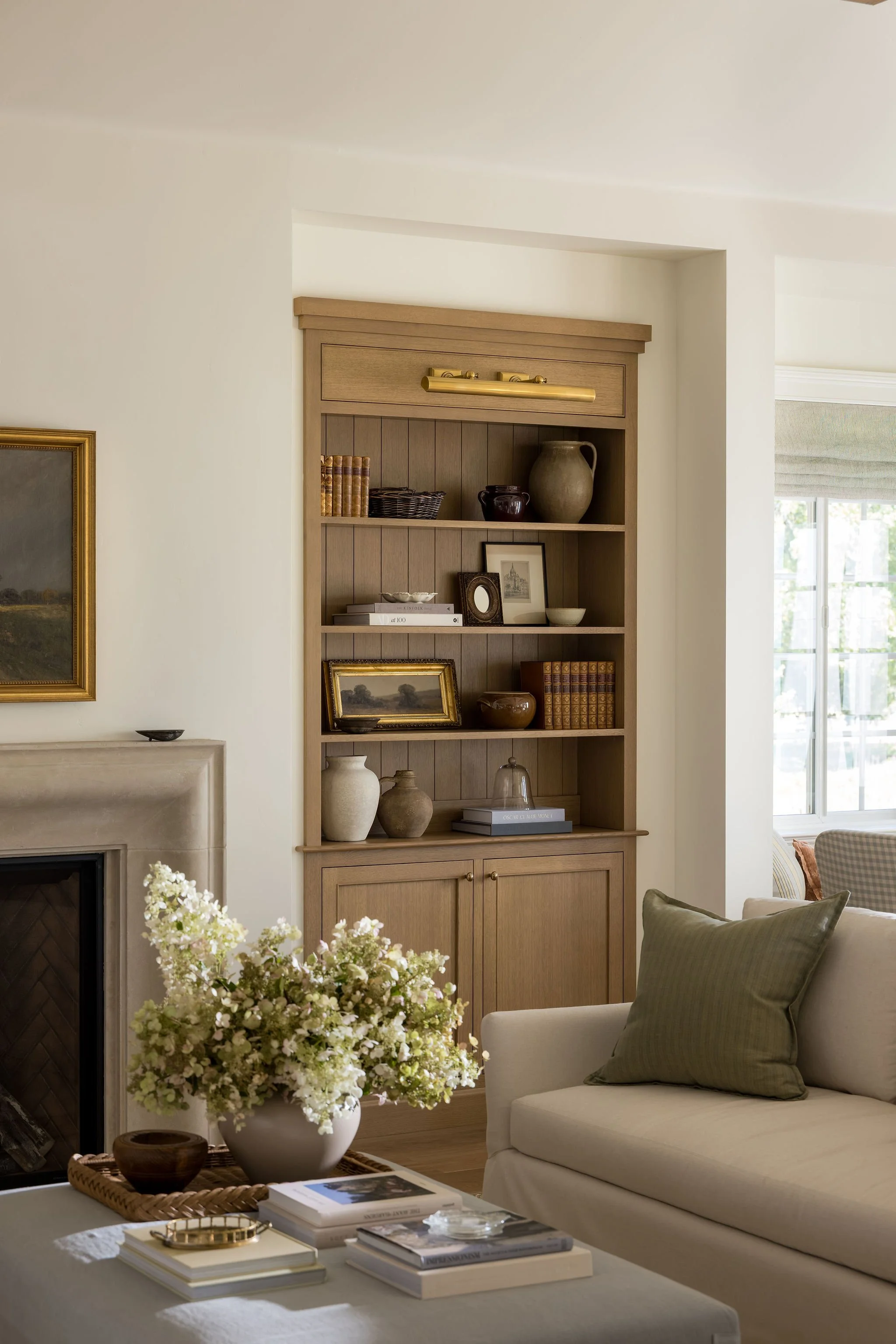 Living room with a beige sofa, a coffee table holding books and decorative items, a large flower arrangement, and a wooden bookshelf with decorative objects and framed photos, illuminated by natural light from large windows.