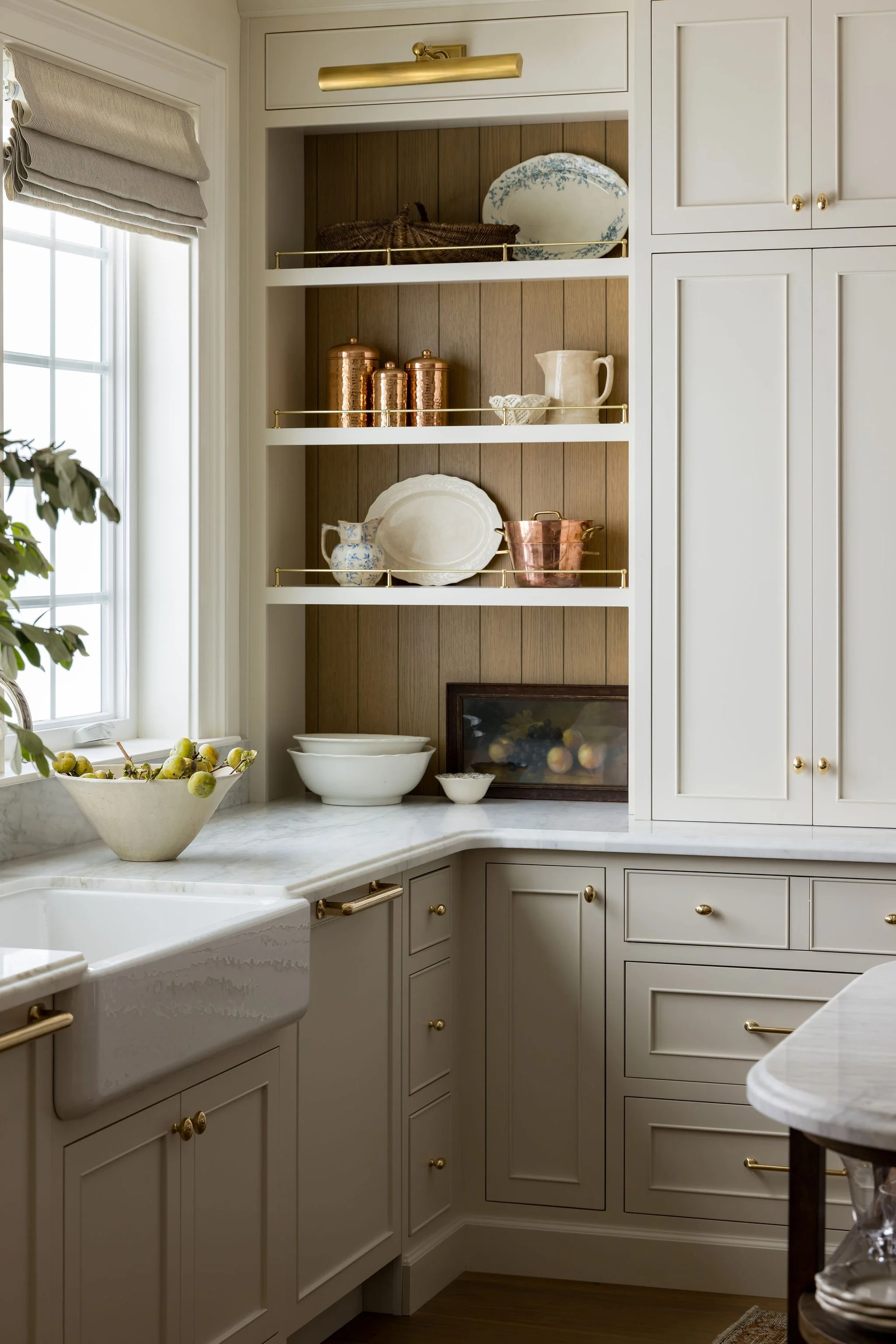 Kitchen with white cabinets, open shelves holding decorative dishes and copper containers, marble countertop, a large window with a beige Roman shade, a bowl of grapes, and a farmhouse sink.