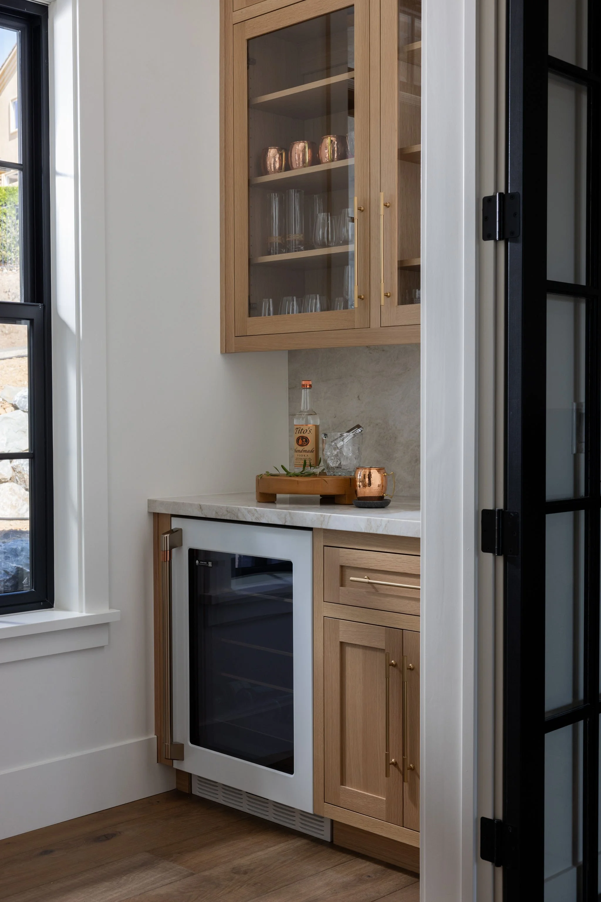 A kitchen area with wooden cabinets, a built-in oven with a black glass door, a countertop with a bottle of Tito's vodka, a glass, and a copper cup. There is a window to the left and a black-framed glass door to the right.