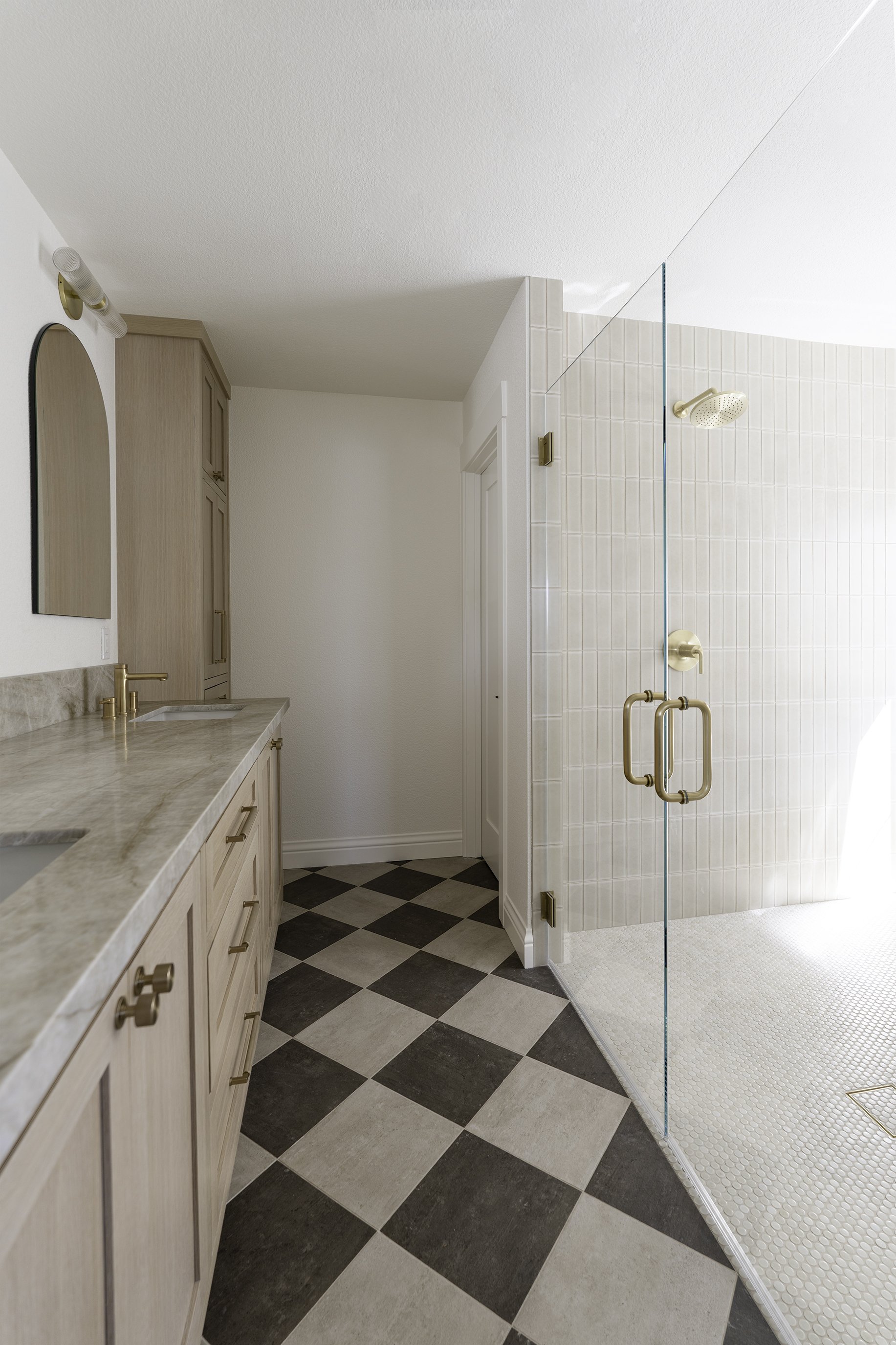 Modern bathroom with a sink vanity, mirror, and a glass-enclosed walk-in shower with gold fixtures. Black and white checkered floor tiles, light-colored cabinetry, and a white door in the background.
