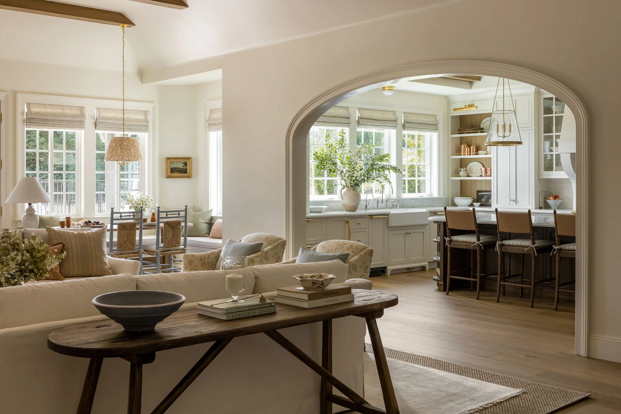 Bright, airy living room with multiple large windows, white cabinetry in the kitchen, beige and cream furniture, and wooden accents including a coffee table and bar stools.