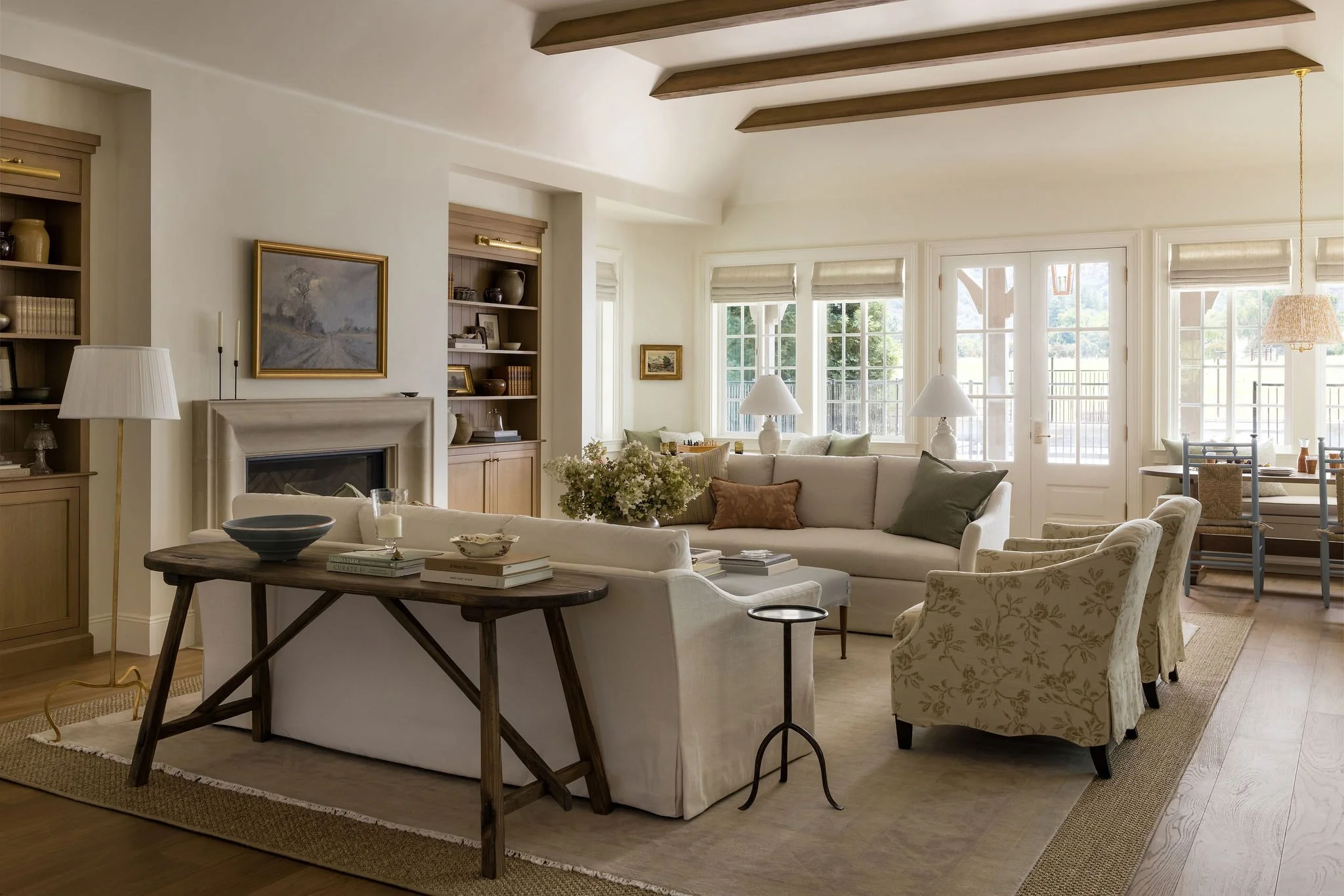A cozy living room with white walls, large windows with blinds, beige and patterned armchairs, a white sofa with throw pillows, a wooden coffee table with books and decor, bookshelves with decorative items, and a fireplace, all illuminated by natural
