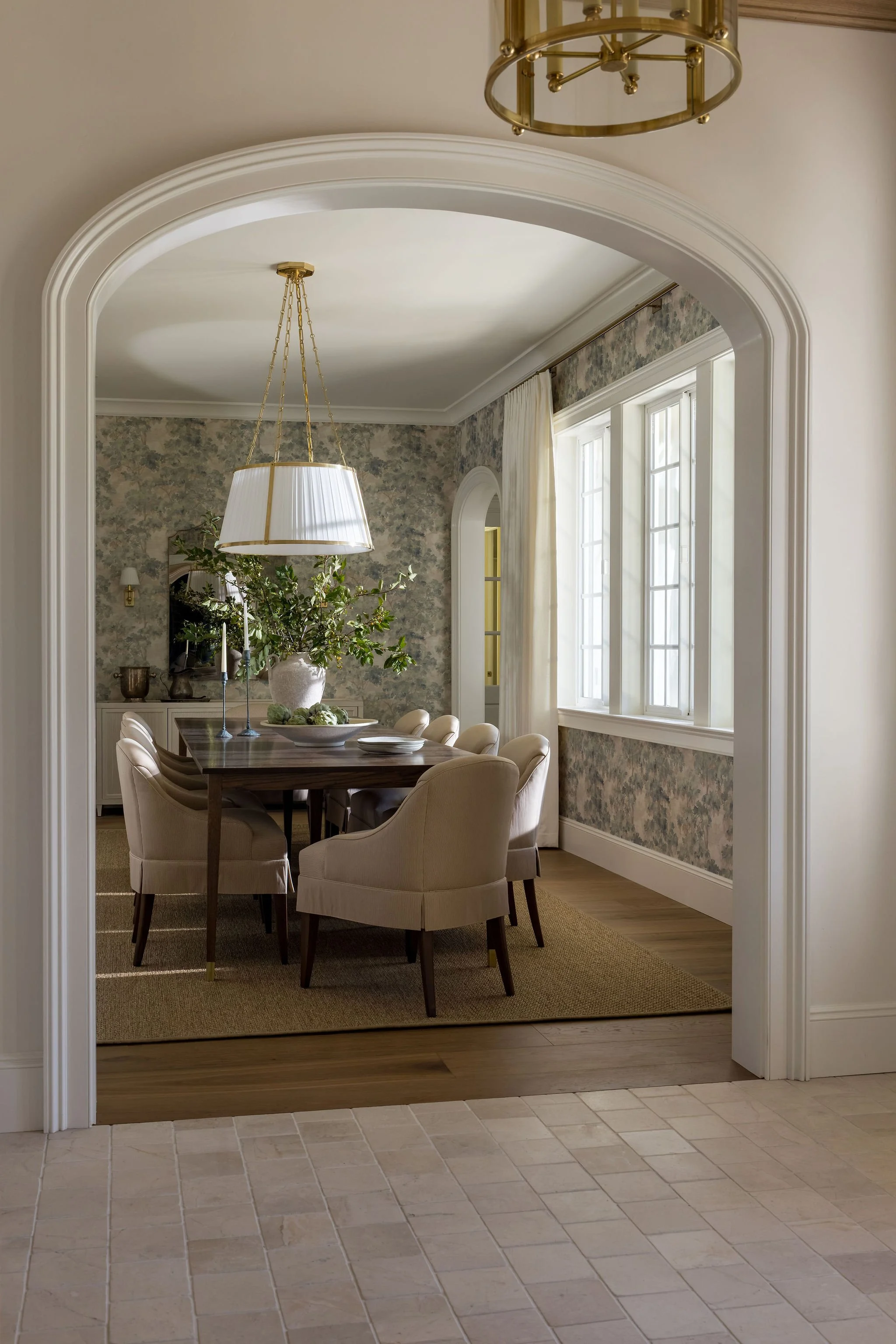 A dining room with a wooden table surrounded by upholstered chairs, a large white pendant light, and a floral wallpapered wall with a window and cream curtains.