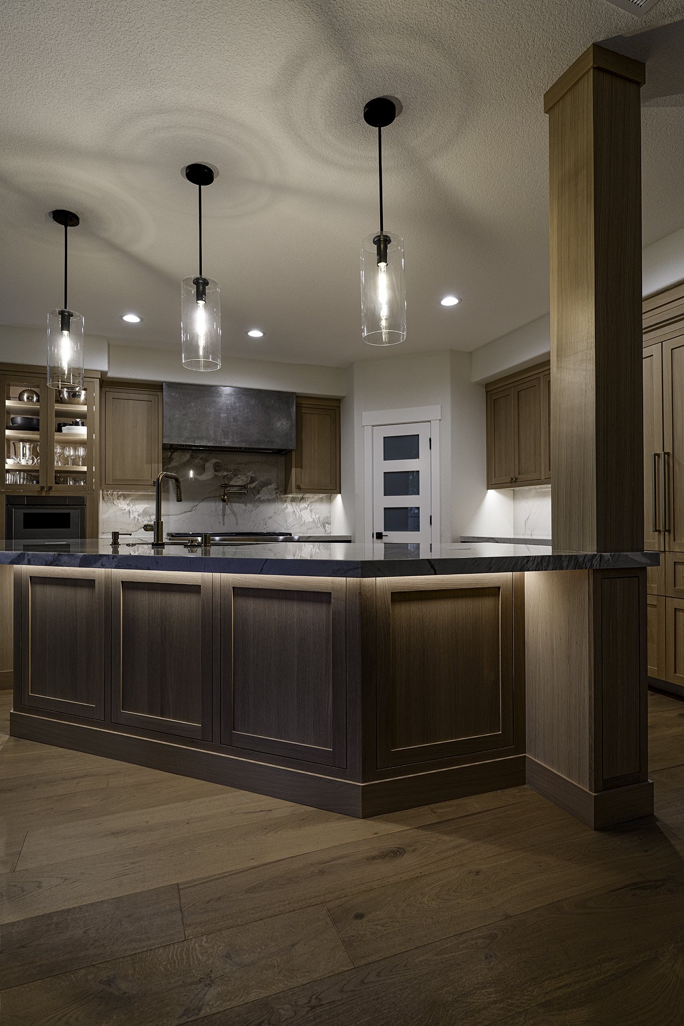 Modern kitchen with wooden cabinets and a dark marble island, pendant lights, and a white door with glass panels.