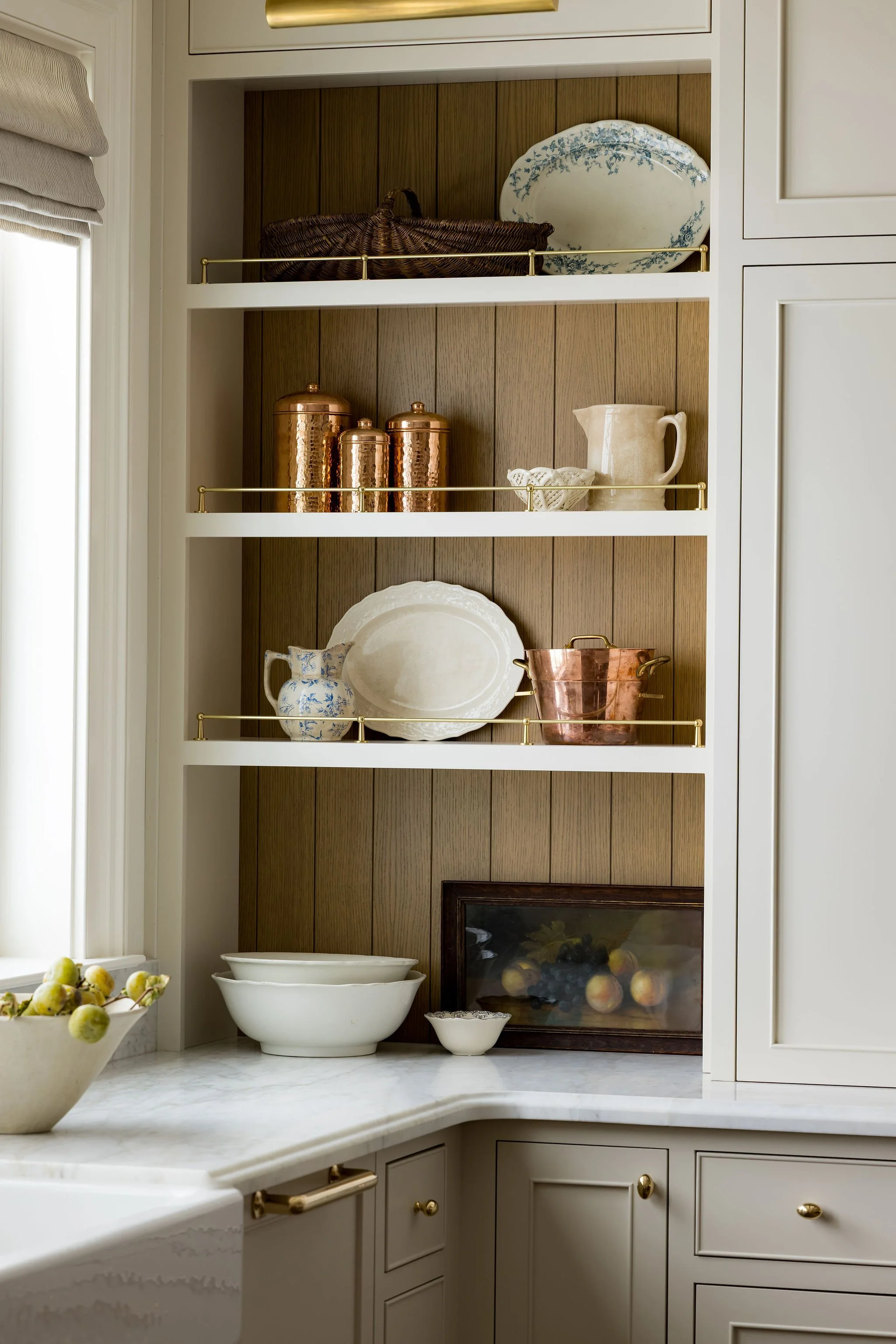 A kitchen cabinet with open shelves displaying decorative dishes, copper containers, and a ceramic pitcher against wood-paneled backs, with additional bowls on the marble countertop and a bowl of green and yellow fruits nearby.