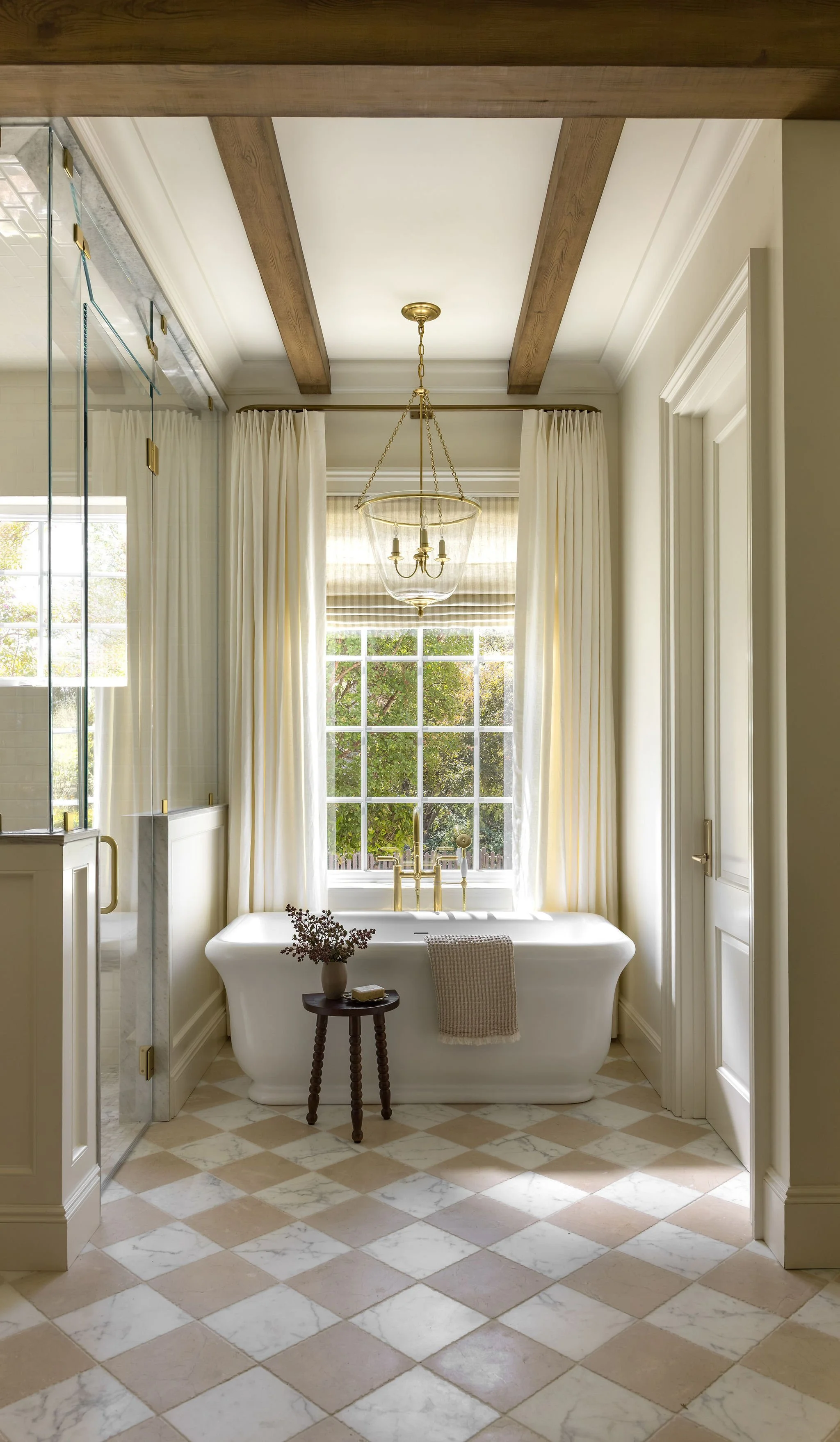 A bathroom with a freestanding bathtub in front of a large window with cream curtains, a chandelier hanging from the ceiling, and a small wooden side table with a vase of flowers and a soap.
