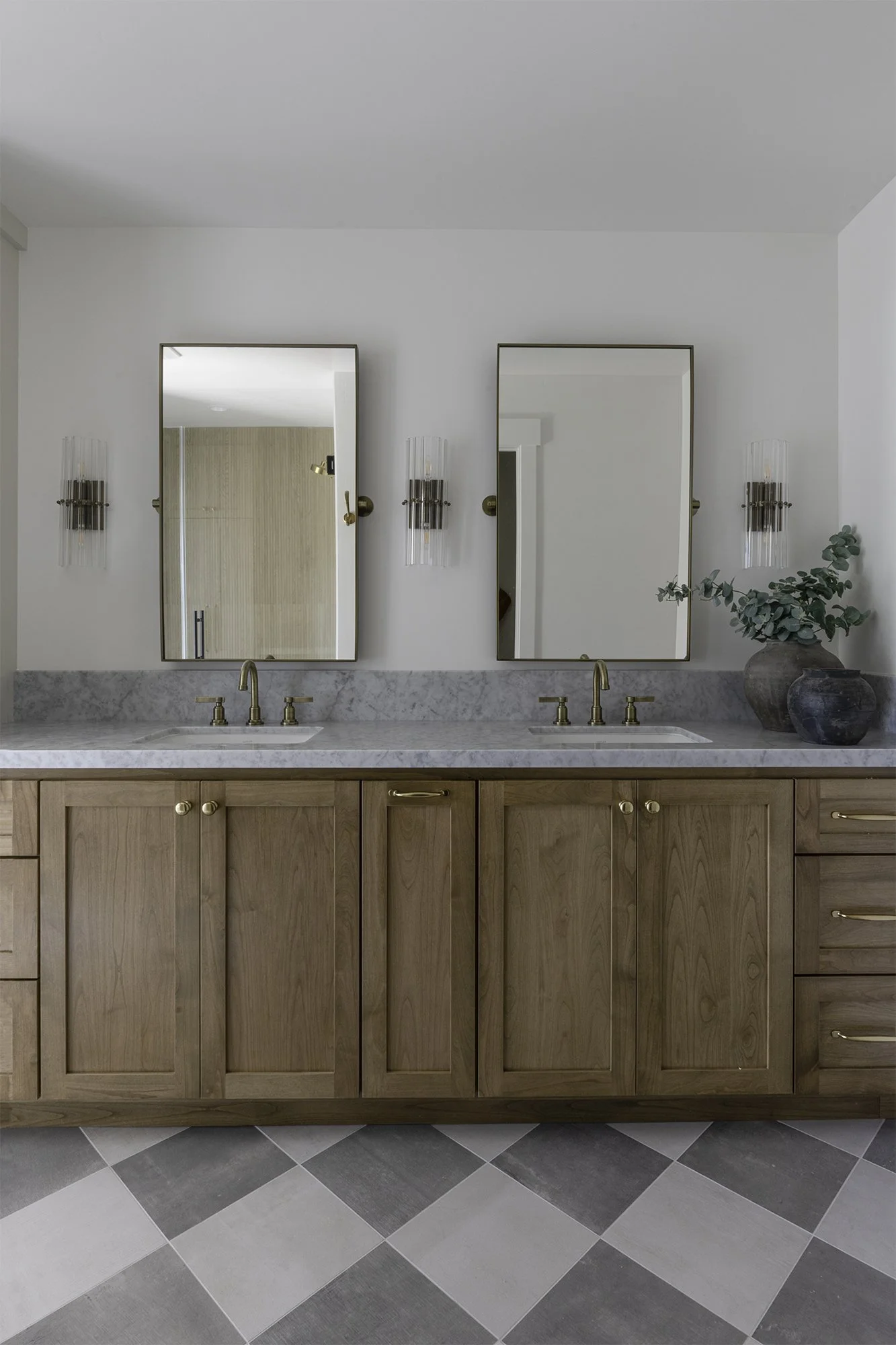 A double bathroom vanity with two mirrors, sconces, a marble countertop, brass faucets, wooden cabinets, and potted plants on the right side.