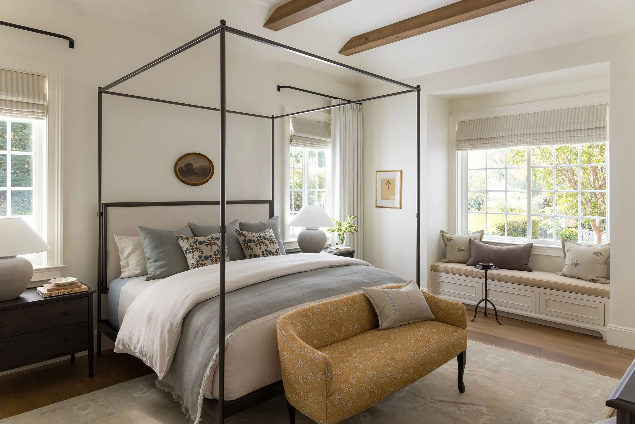 A bedroom with a four-poster bed, nightstand, and a small window seat with pillows. The room has large windows with blinds, light-colored walls and ceiling, and wooden beams. Decor includes framed pictures and a lamp on the nightstand.
