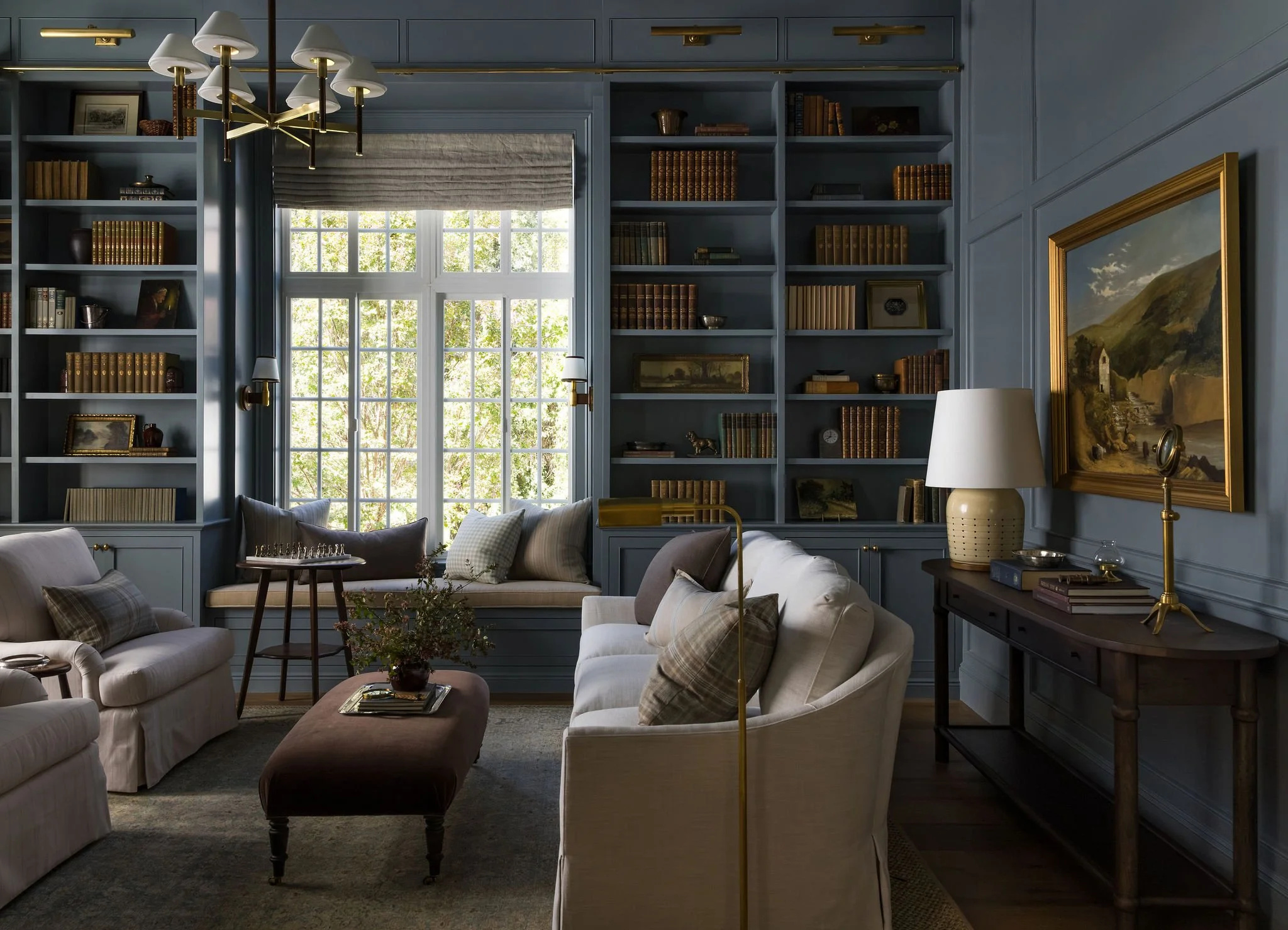 Living room with built-in blue bookshelves, large window with blinds, beige cushioned armchairs, a brown ottoman, a white lamp, and artwork on the walls.
