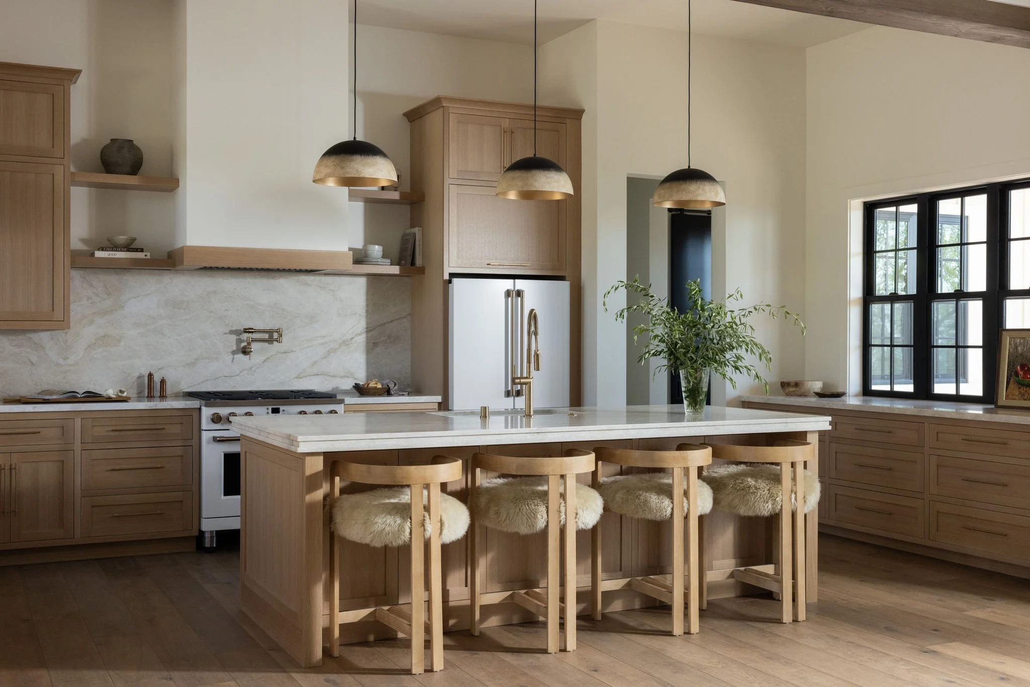 A modern kitchen with wooden cabinetry, a large central island with a marble countertop, four wooden bar stools with plush sheepskin cushions, pendant lights hanging above the island, a potted plant near a window, and a stainless steel refrigerator.