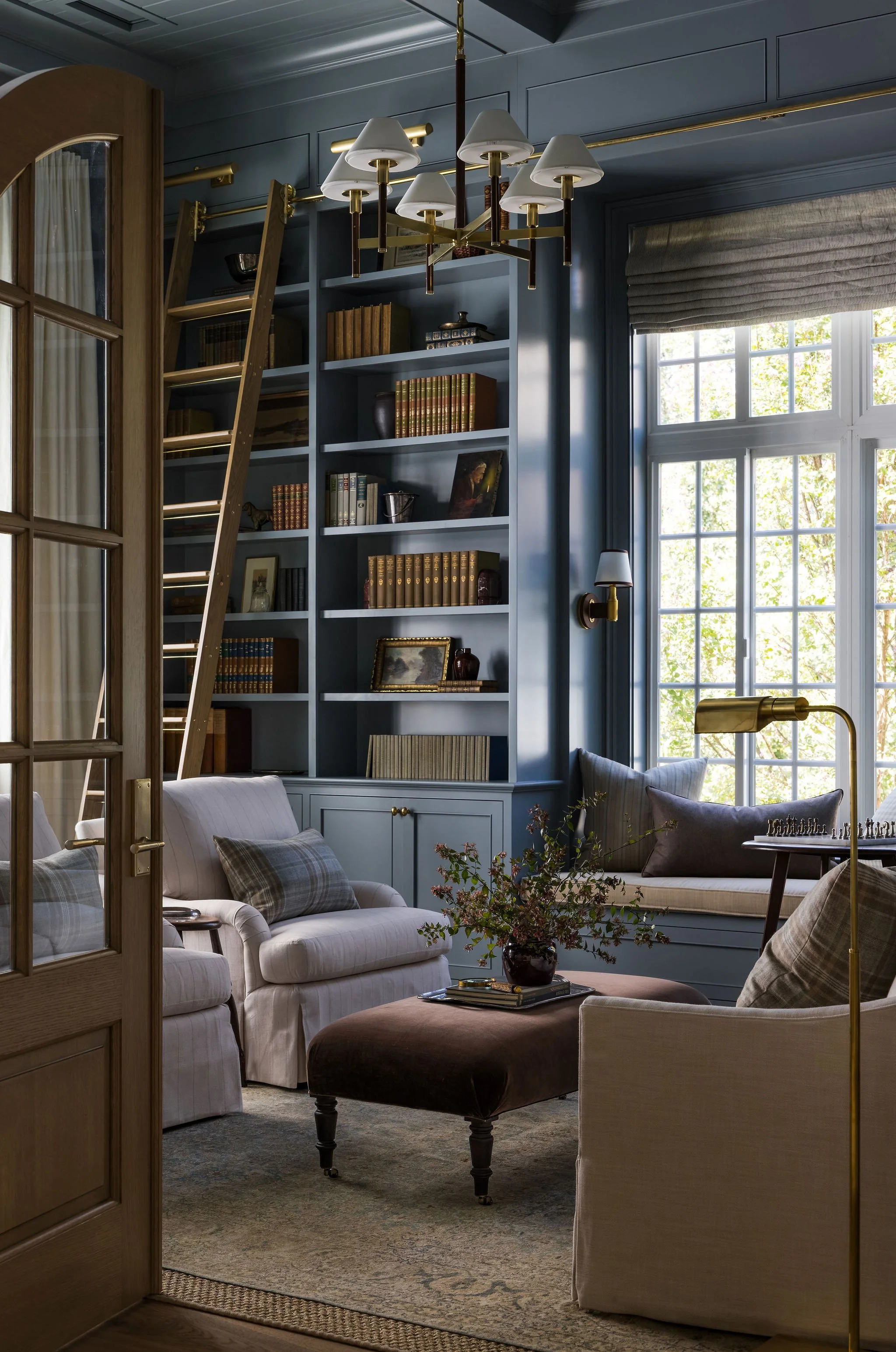 A cozy living room with a blue built-in bookshelf filled with books and decor, a large window with blinds, white and beige sofas with plaid and solid pillows, a dark brown ottoman with a flower arrangement, and a brass and white chandelier overhead.