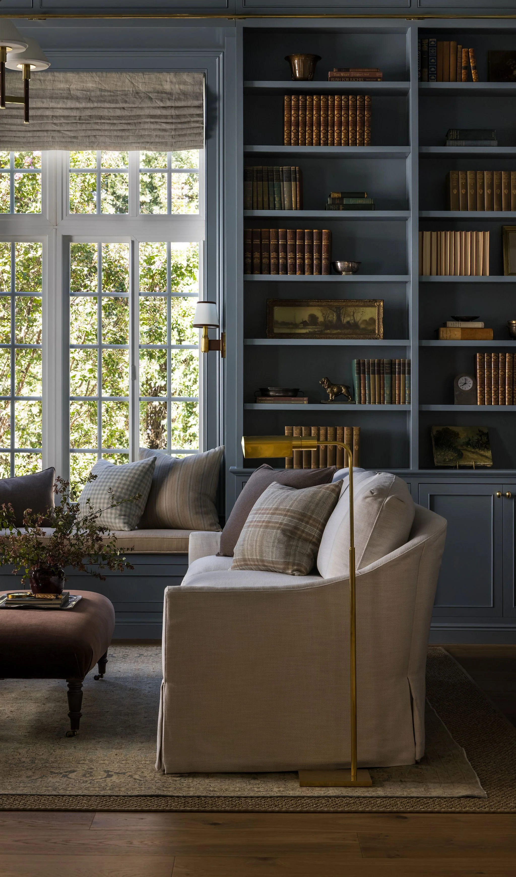A cozy living room corner with a large window, beige armchair with pillows, a dark brown ottoman table with flowers, a blue built-in bookshelf filled with books and decorative items, and a brass floor lamp.