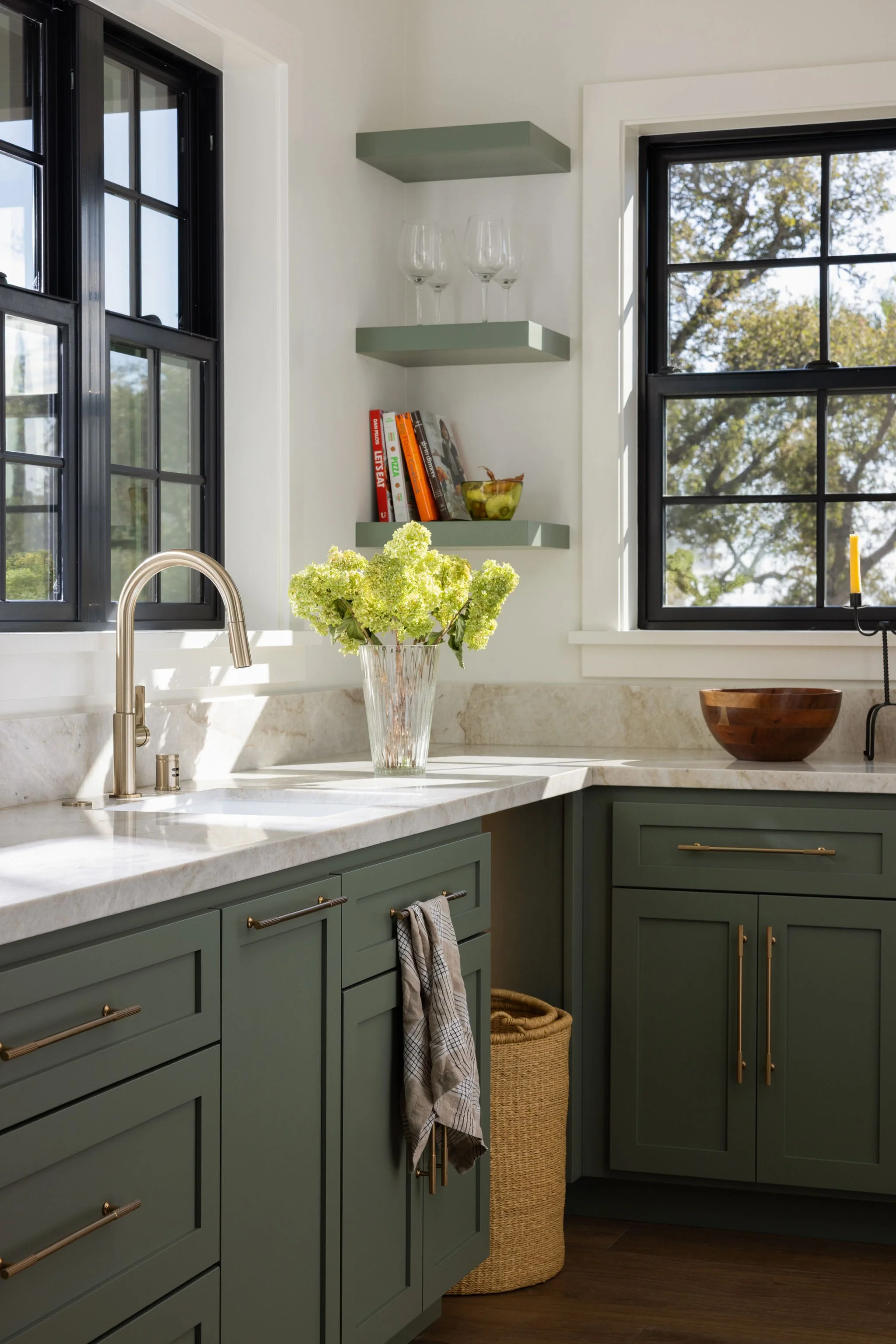 Kitchen with green cabinets, marble countertop, black window frames, a vase with green hydrangeas, and a paper towel holder.