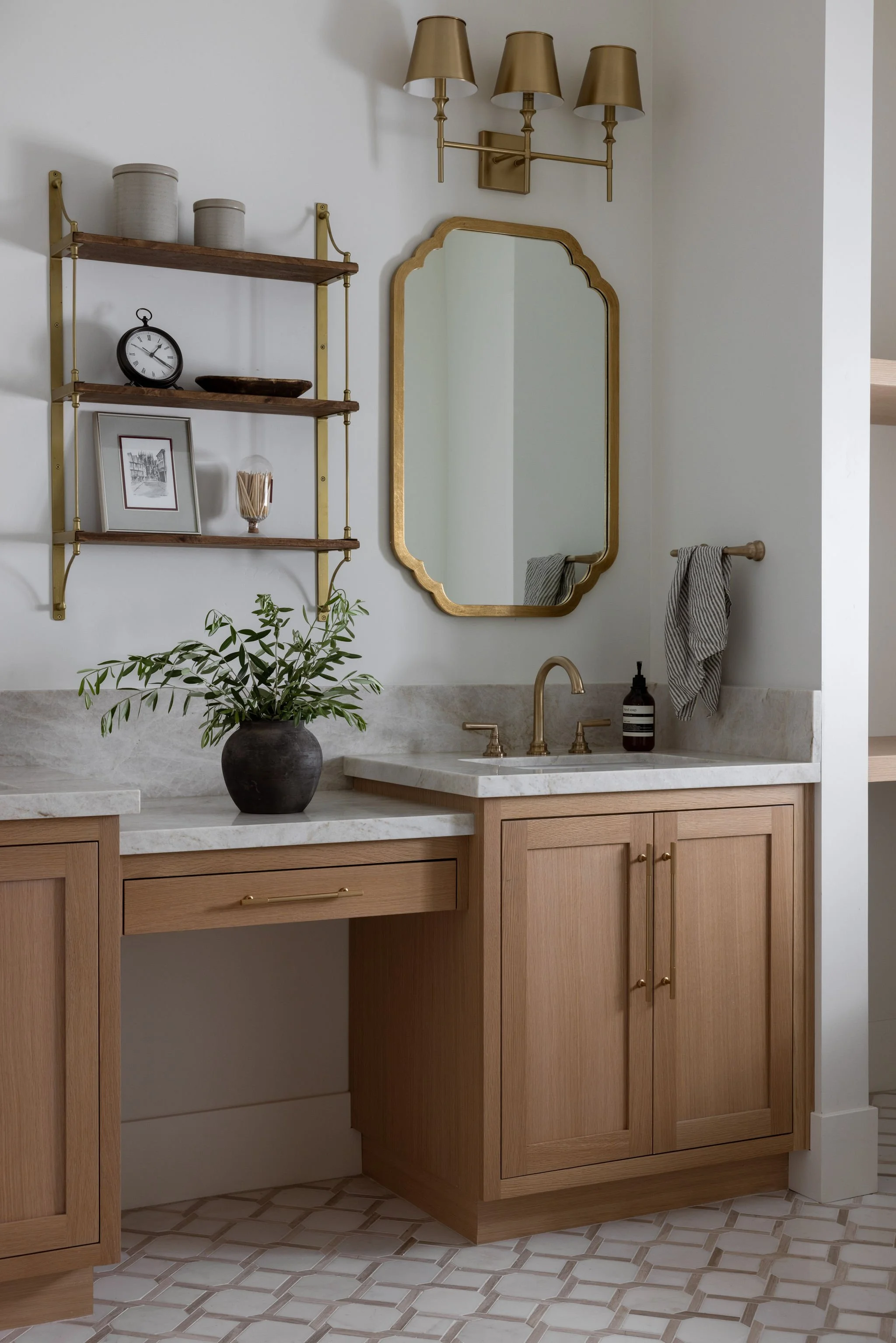 A bathroom vanity with a marble countertop, wooden cabinets, a brass faucet, and a mirror with a gold frame. There is a potted plant on the counter, a black soap dispenser, and a hand towel hanging on a towel rod. Above, a gold light fixture with thr
