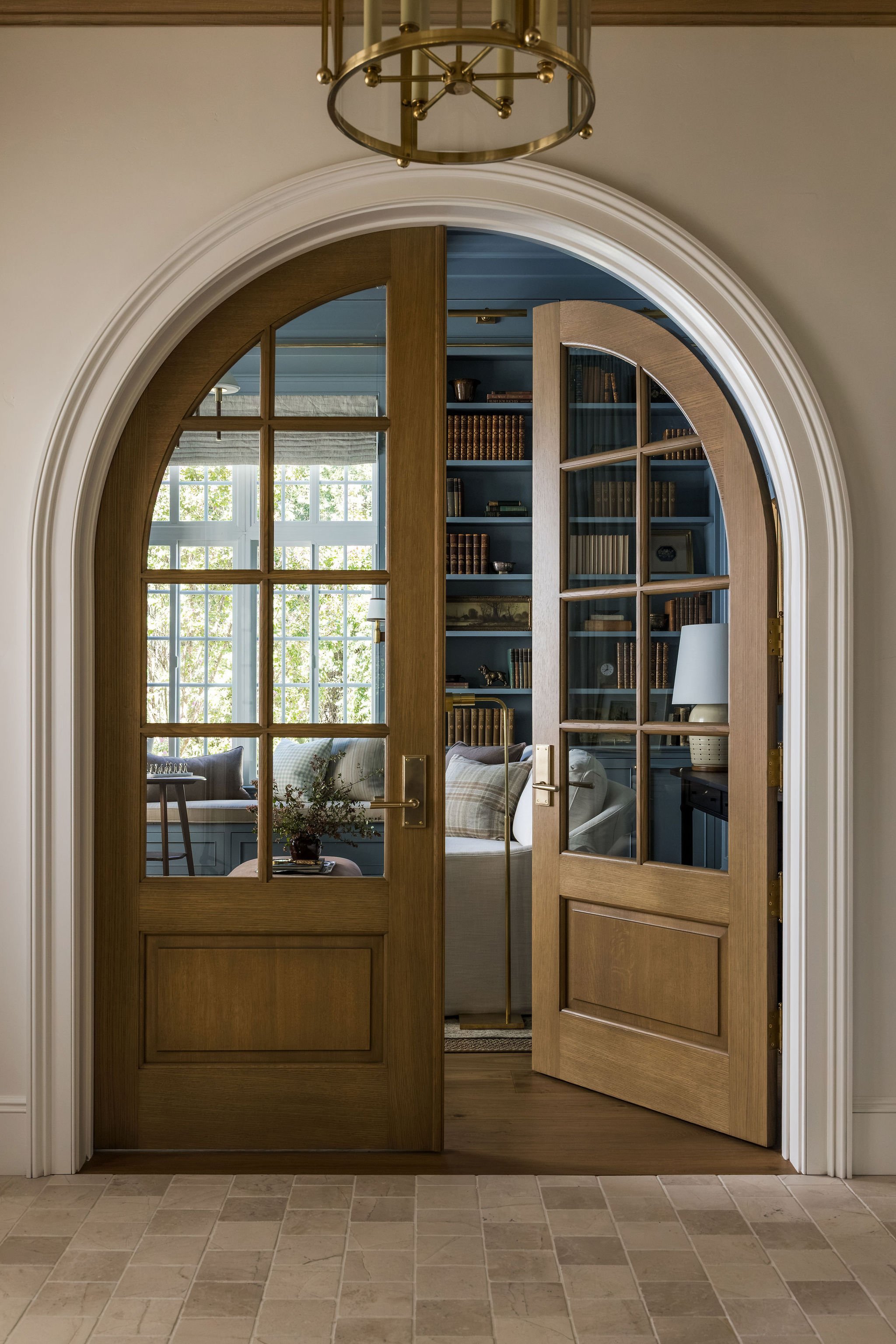 Open wooden double doors with glass panes in an entryway, revealing a living room with a large window, bookshelf, and furniture inside.