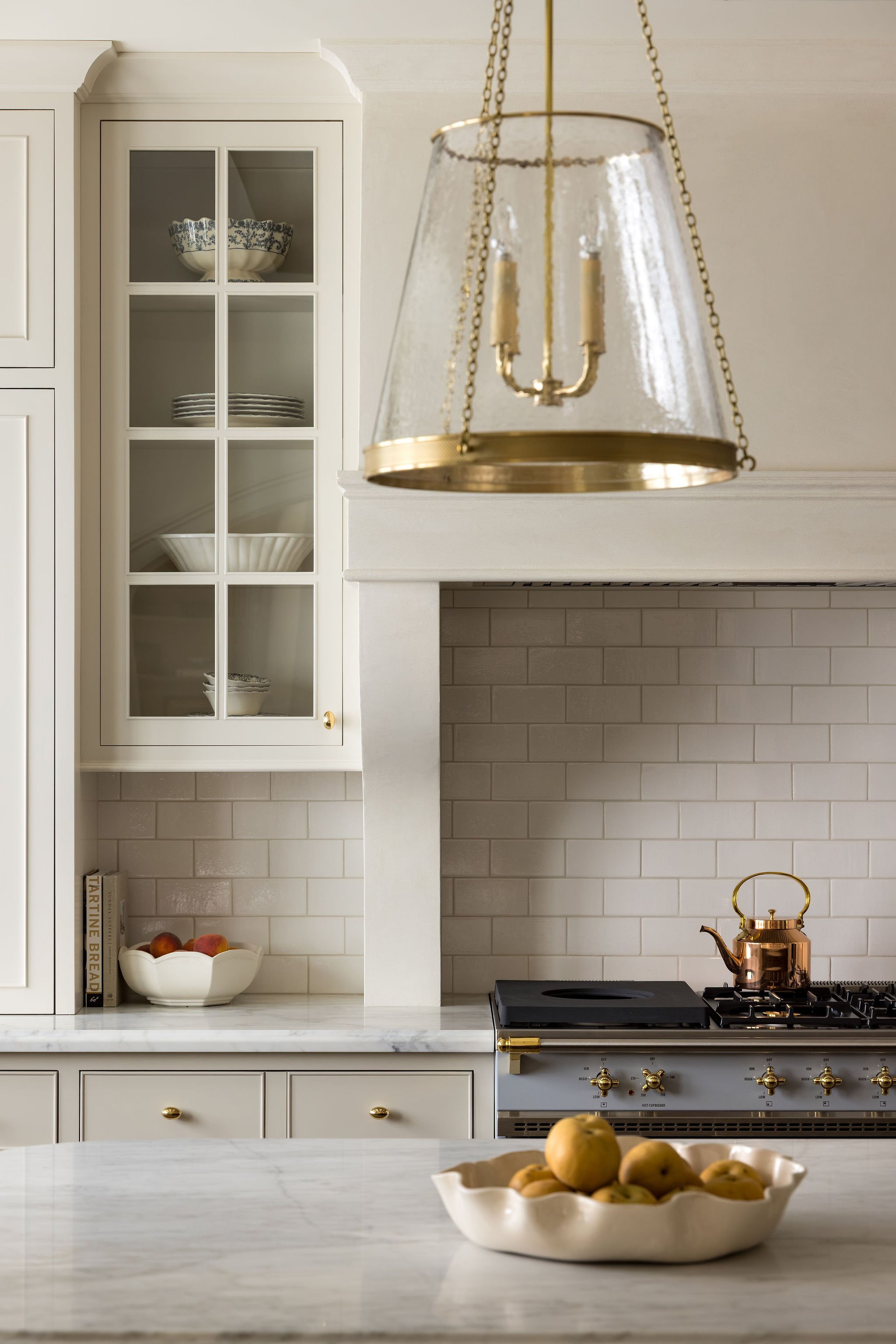 A kitchen scene with a white marble countertop, a white ceramic fruit bowl filled with apples, a stove with a copper kettle, and a white built-in cabinet with glass doors displaying dishes and bowls. A gold-colored chandelier hangs from the ceiling.