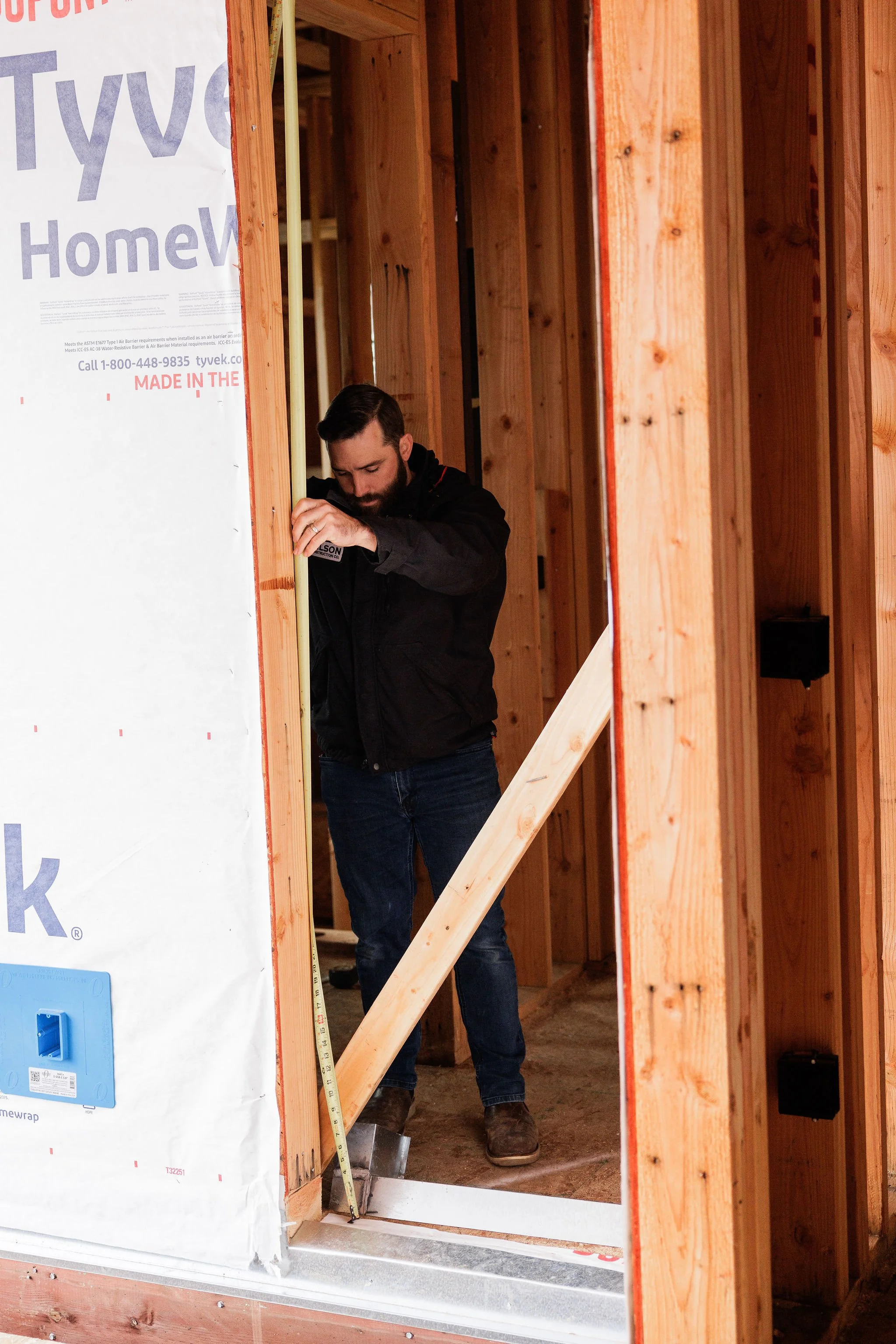 A man working inside a wooden frame structure of a house under construction, measuring with a tape measure.