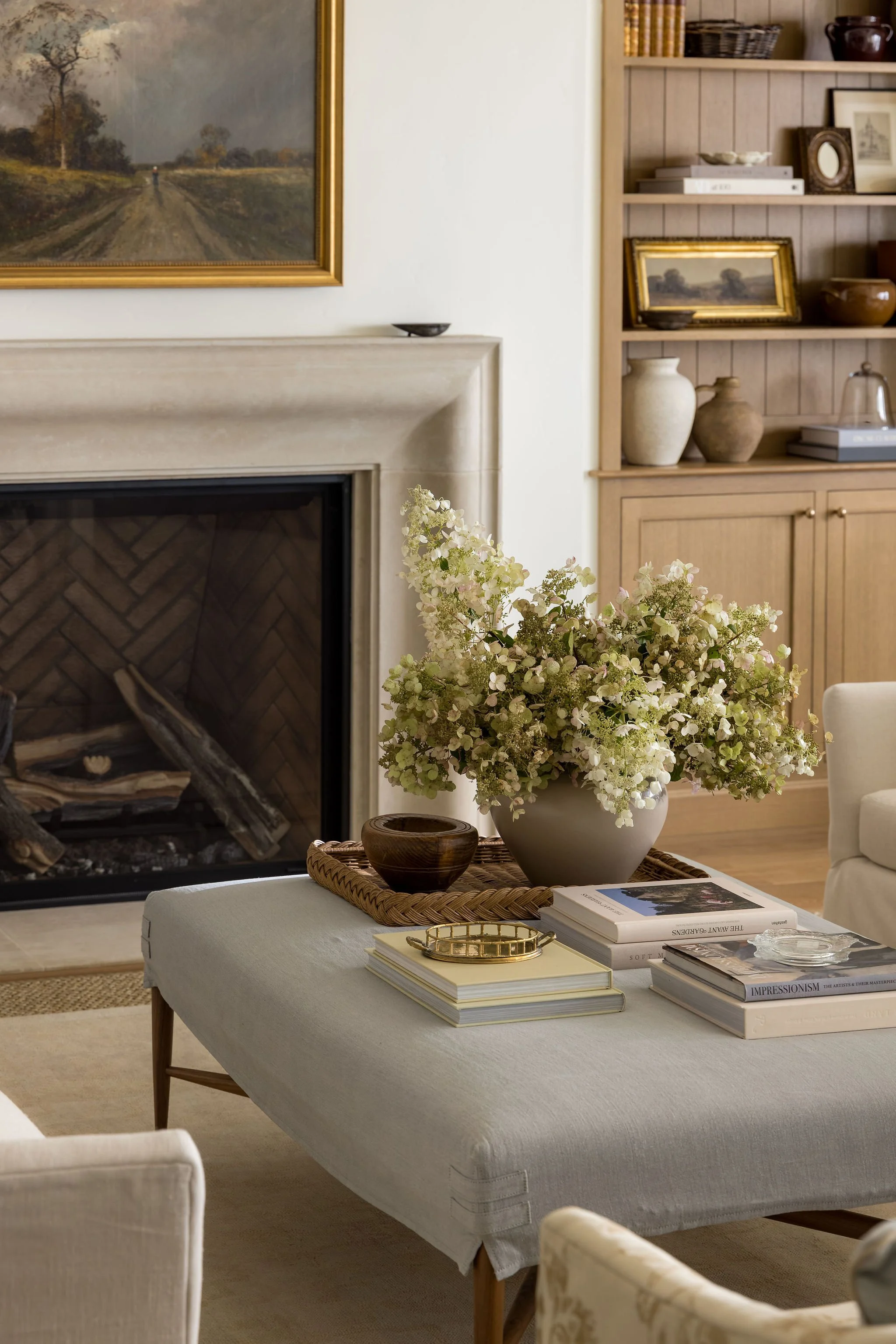 A living room with a white couch and a table holding a large vase of white and light green flowers, several books, and decorative items. In the background, a fireplace with a painting above it and a wooden bookshelf with decorative items and framed p