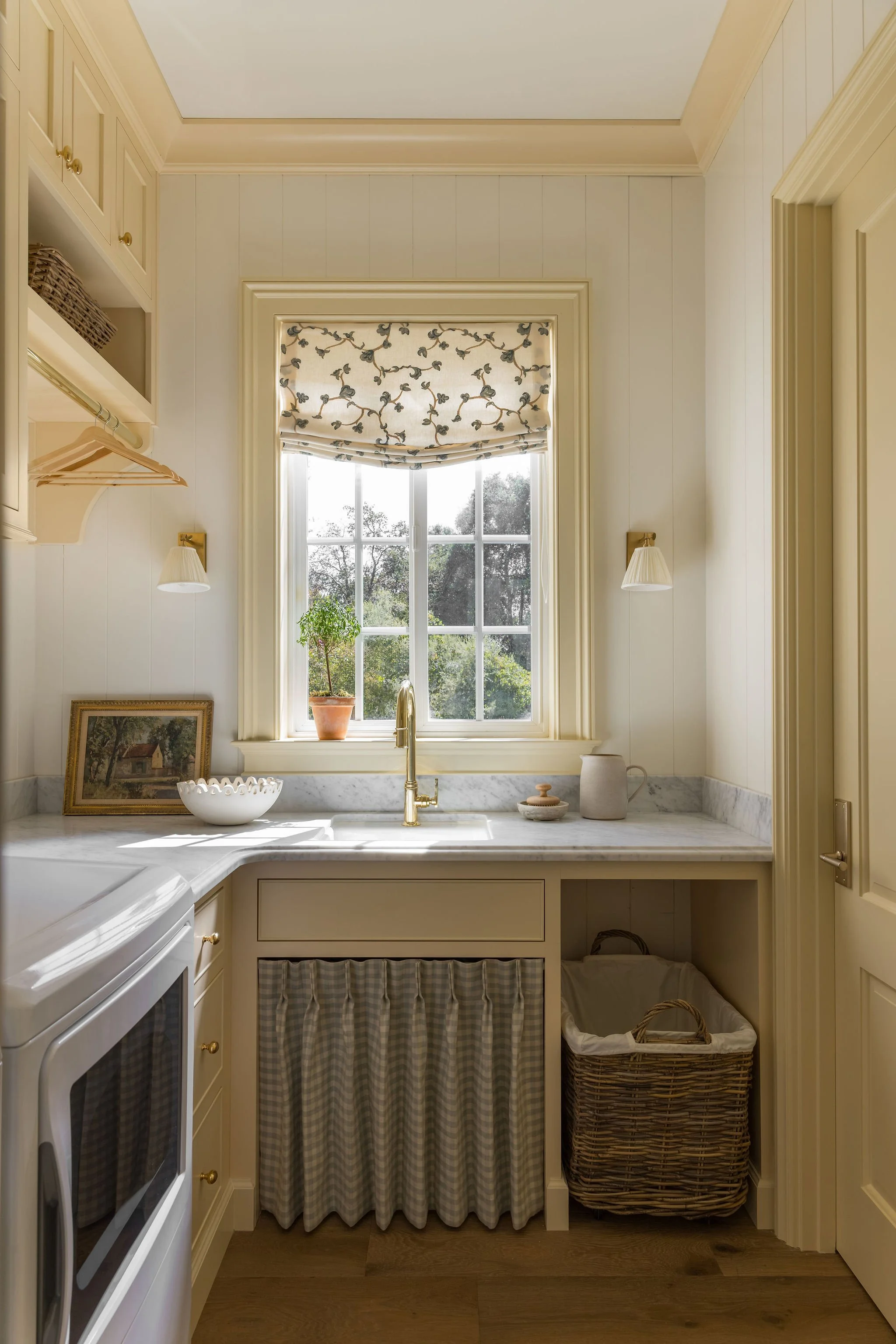A cozy kitchen with cream-colored cabinets and a window with floral Roman shade. The window sill has a potted plant, and there are wall-mounted sconces with fabric shades. On the counter, there is a picture frame, a bowl, and a pitcher. Below the win
