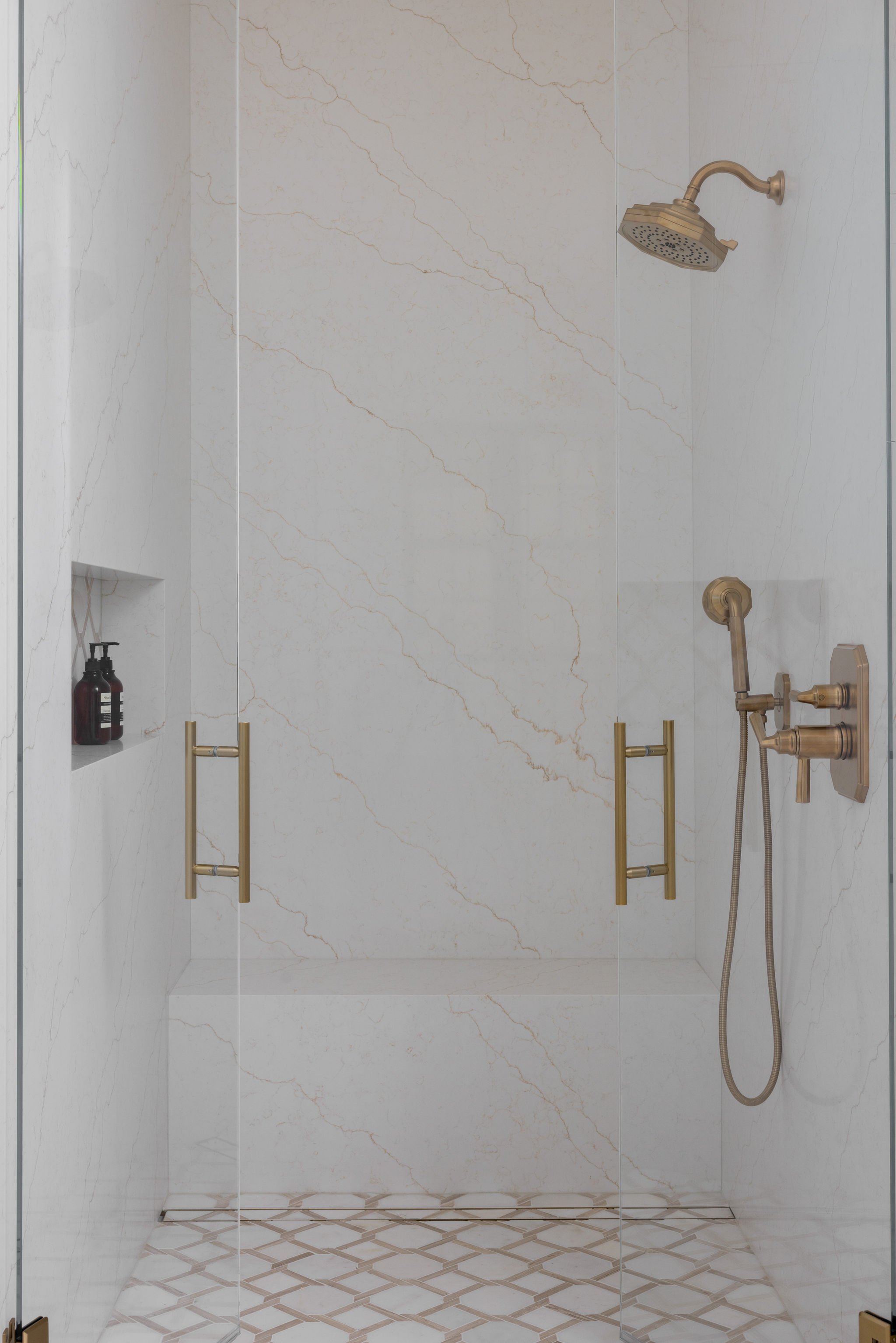 A walk-in shower with white marble walls with gold veining, a gold showerhead, and a handheld spray. There are two gold towel bars and a built-in shelf with two brown bottles.