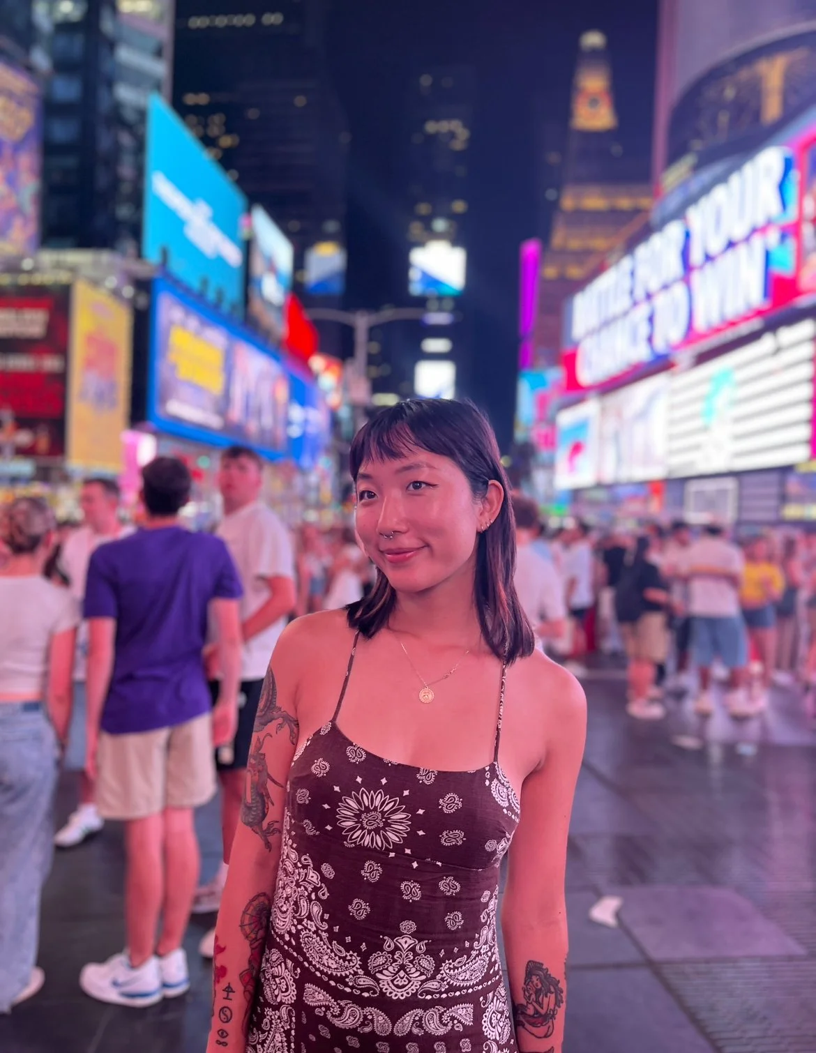 A young woman with tattoos on her arms and a nose ring, wearing a dark sundress with a white paisley pattern, standing in Times Square at night with bright neon signs and a crowd of people in the background.