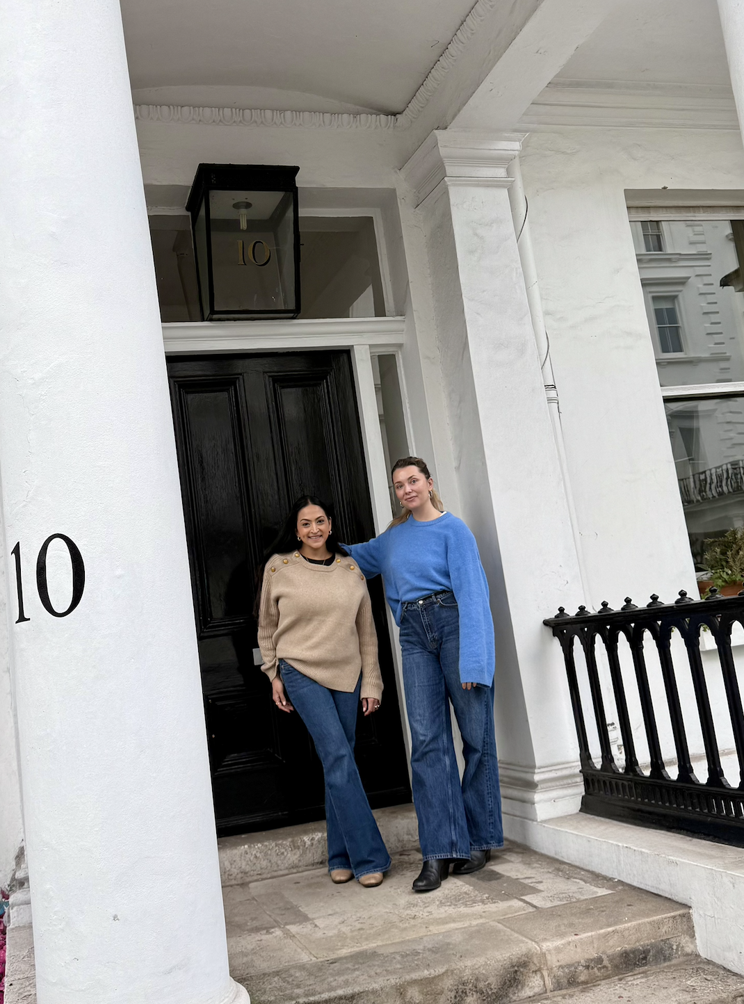 Two women standing on the front steps of a white house with the number 10 on a pillar, black front door, and large windows, posing for a photo.