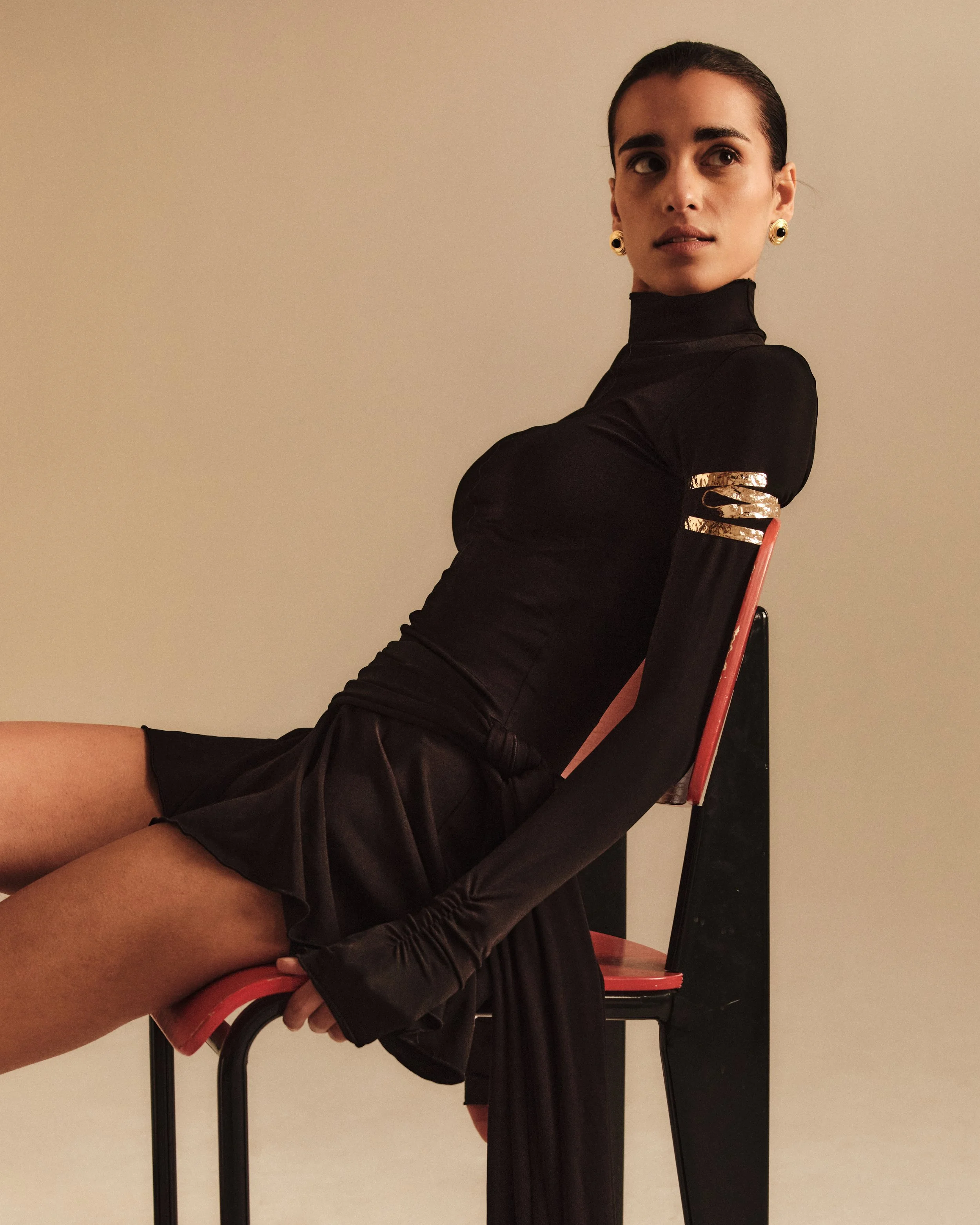 A woman sitting sideways on a black and red chair, wearing a black long-sleeve top with gold accents and the same color skirt, with her hair slicked back, looking to her right.