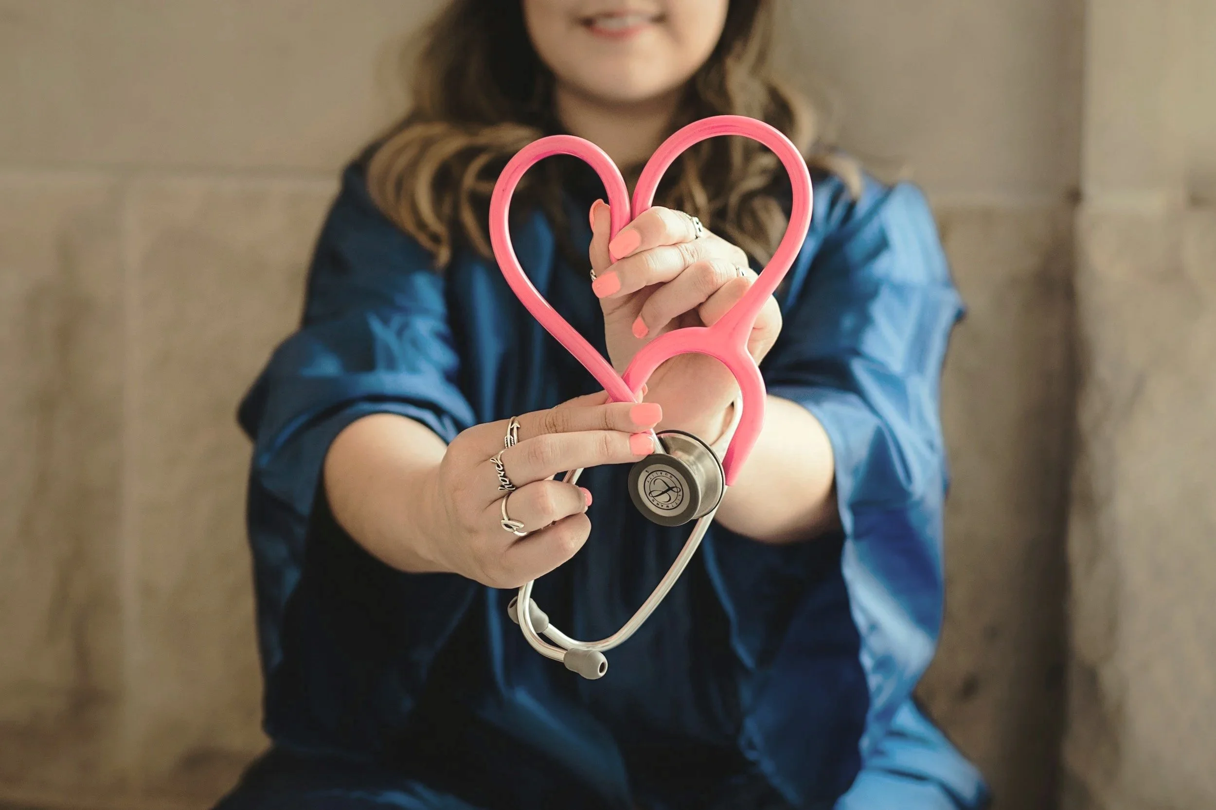 A Doctor holding a pink heart-shaped stethoscope, smiling. They have long hair, wear rings and a blue jacket.