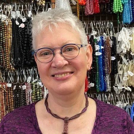 A smiling woman with short white hair, glasses, and earrings in a jewelry store surrounded by colorful beads and necklaces.