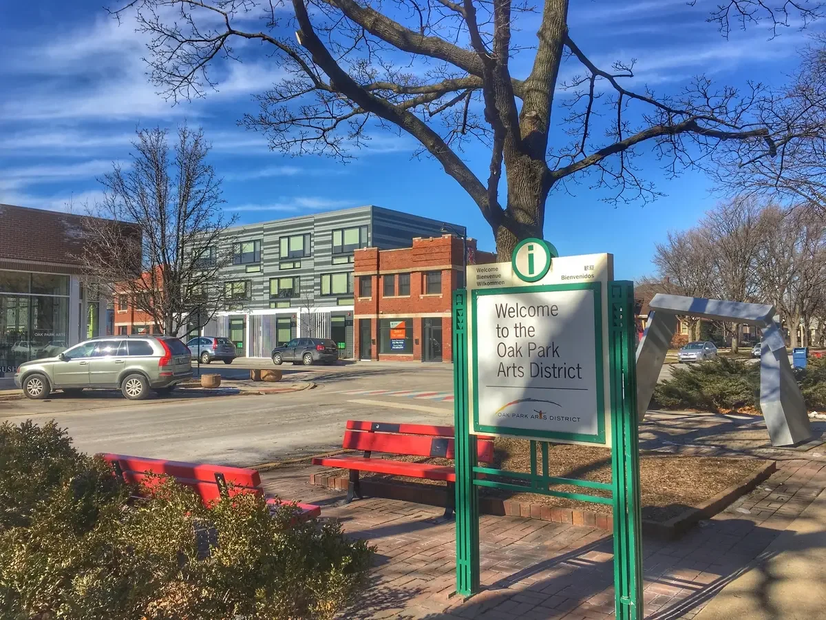 Street scene in the Oak Park Arts District with a sign welcoming visitors, parked cars, a leafless tree, benches, and buildings under a blue sky with wispy clouds.