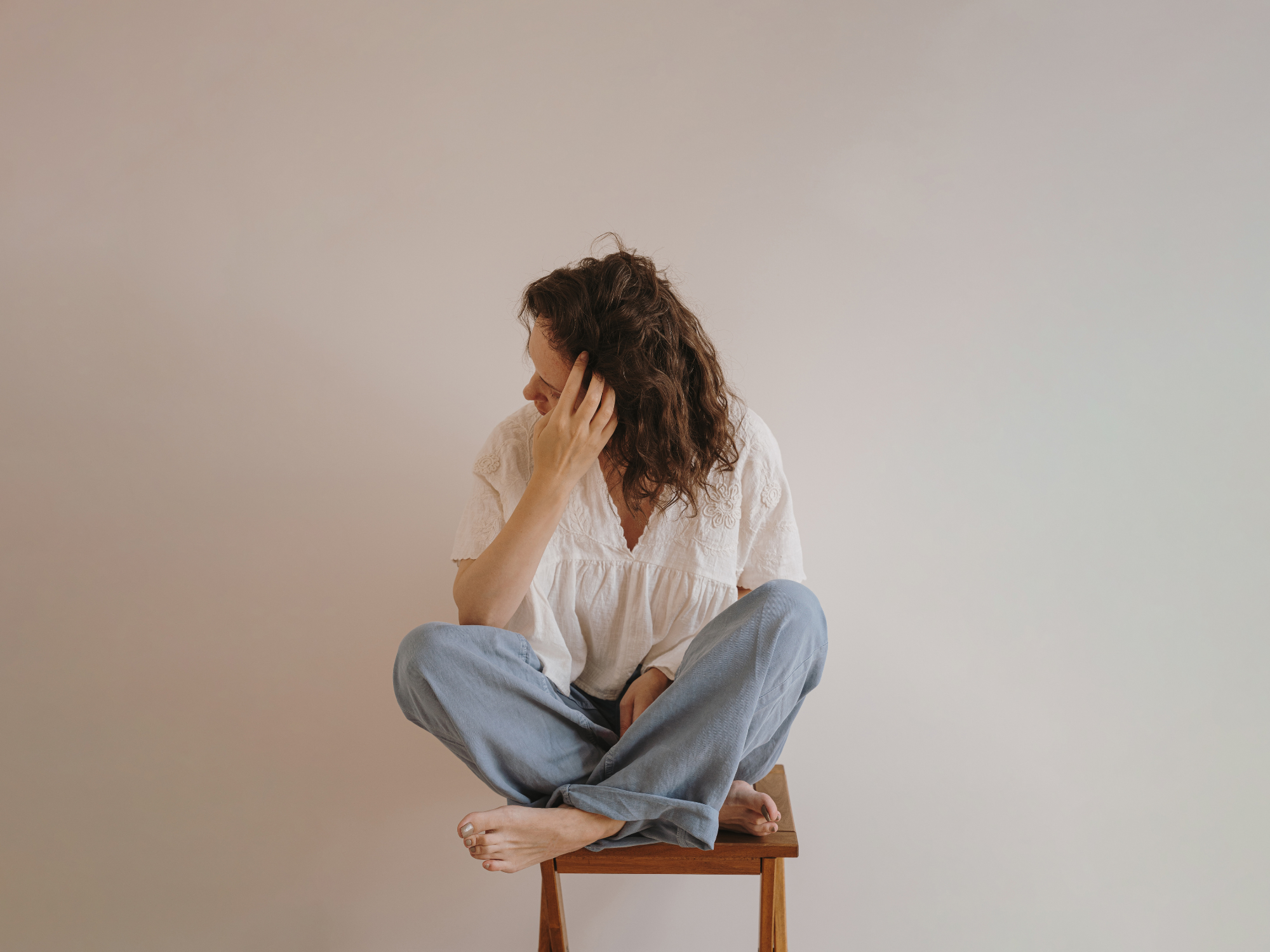 Une femme assise en indien sur une chaise en bois, touchant son oreille, regardant vers le bas, vêtue d'une chemise blanche et de jeans