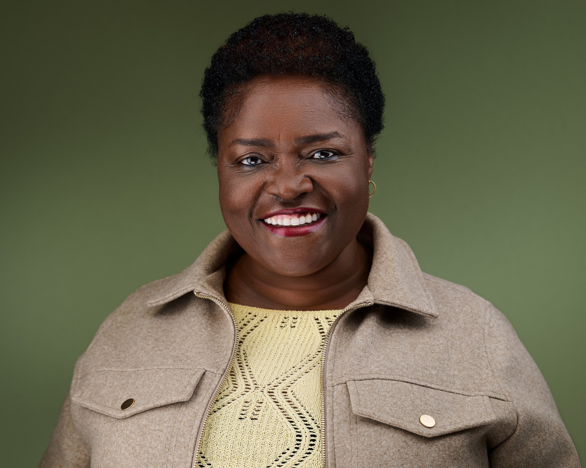 Vivica smiling with dark hair and dark skin, wearing a beige jacket over a yellow knitted top, standing against a green background.