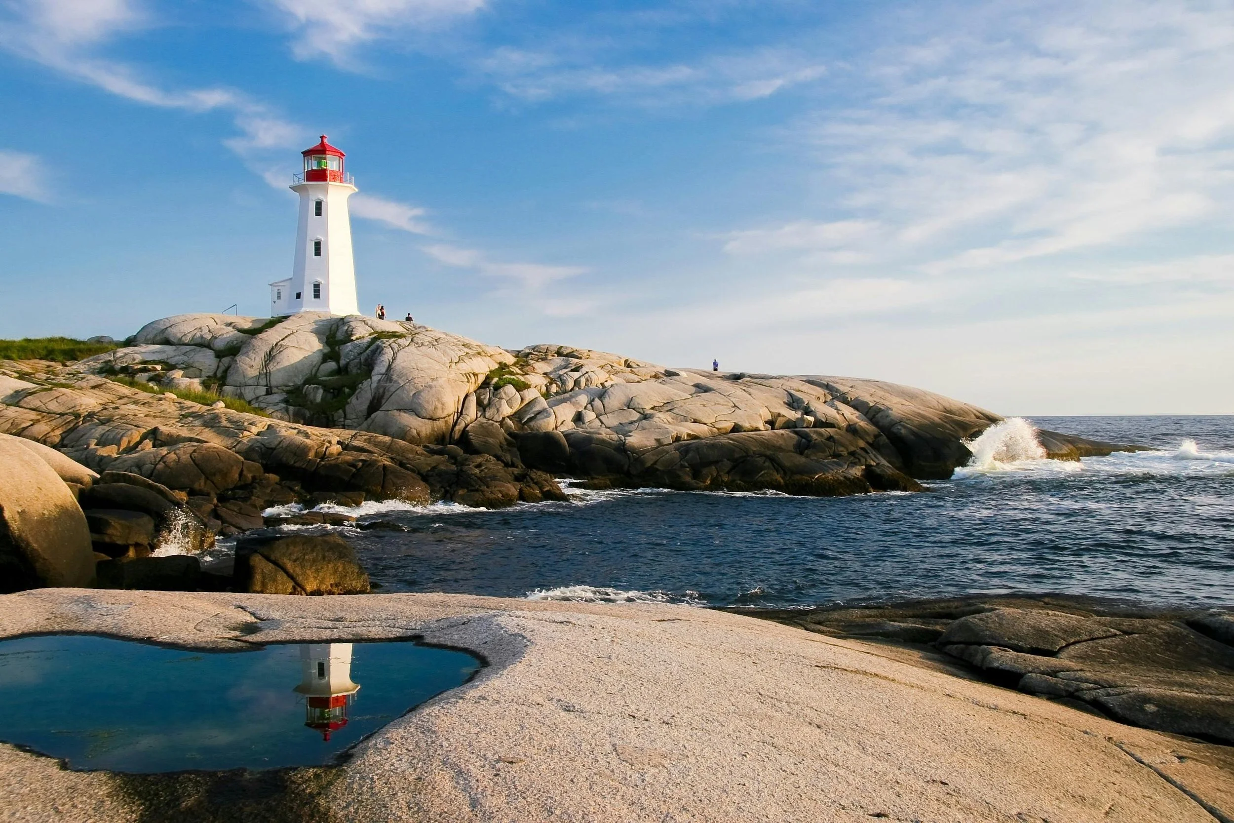 A white lighthouse with a red top on rocky coastline, ocean waves crashing nearby, blue sky with some clouds, and a small water pool reflecting the lighthouse in the foreground.