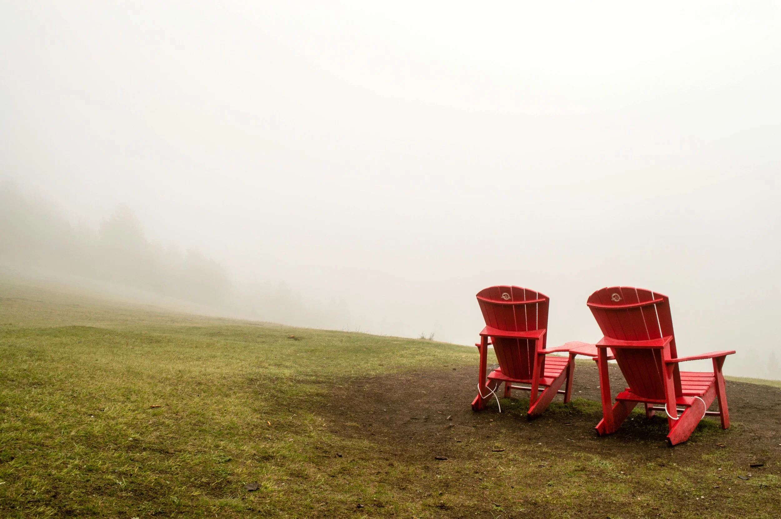 Two red Adirondack chairs on a grassy hilltop in foggy weather with a white, misty background.