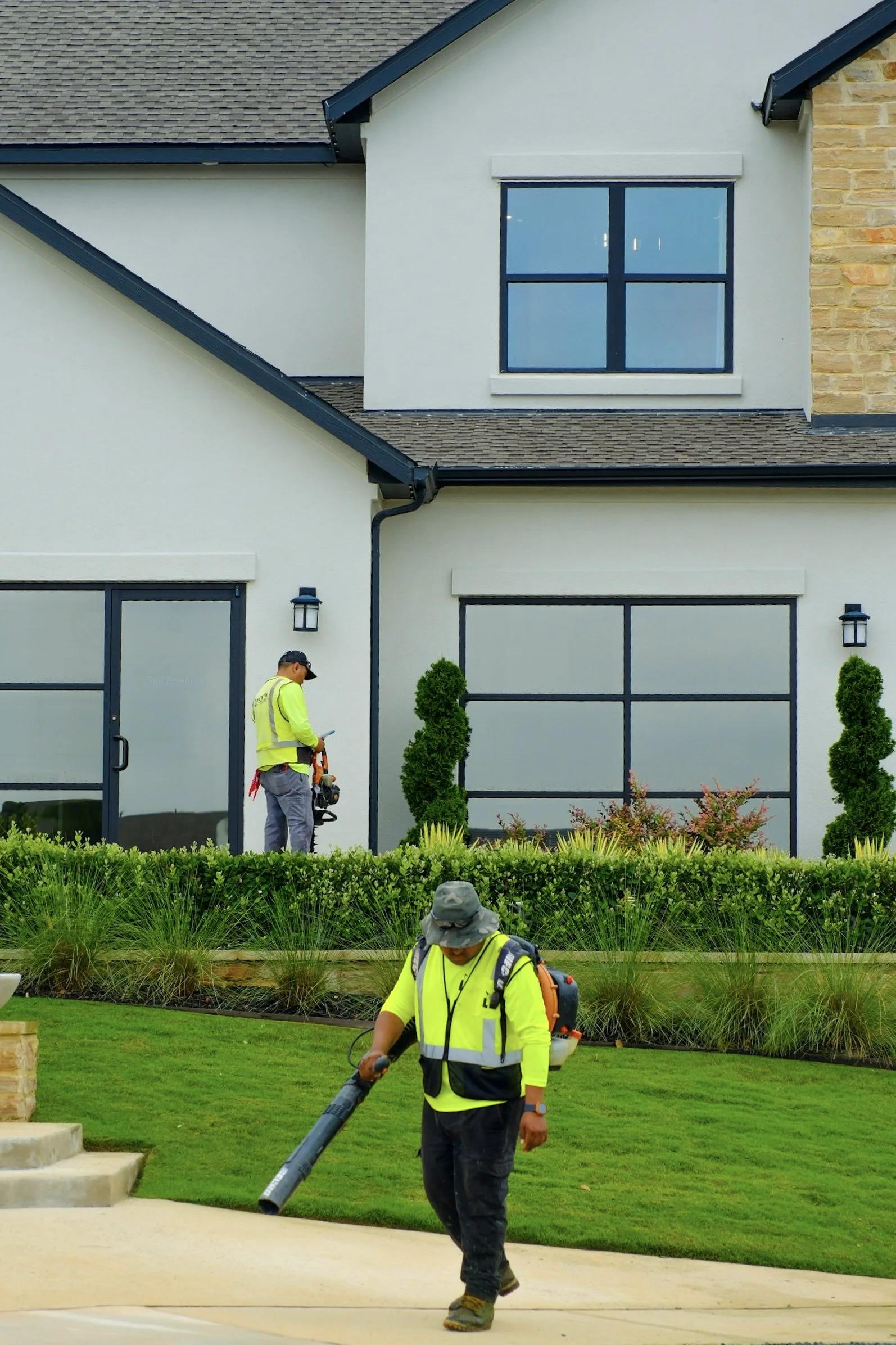 Two workers in neon yellow safety shirts and broad-brim hats working outside a house; one is leaf blowing on a concrete sidewalk, while the other is using a leaf blower near the house's garden.