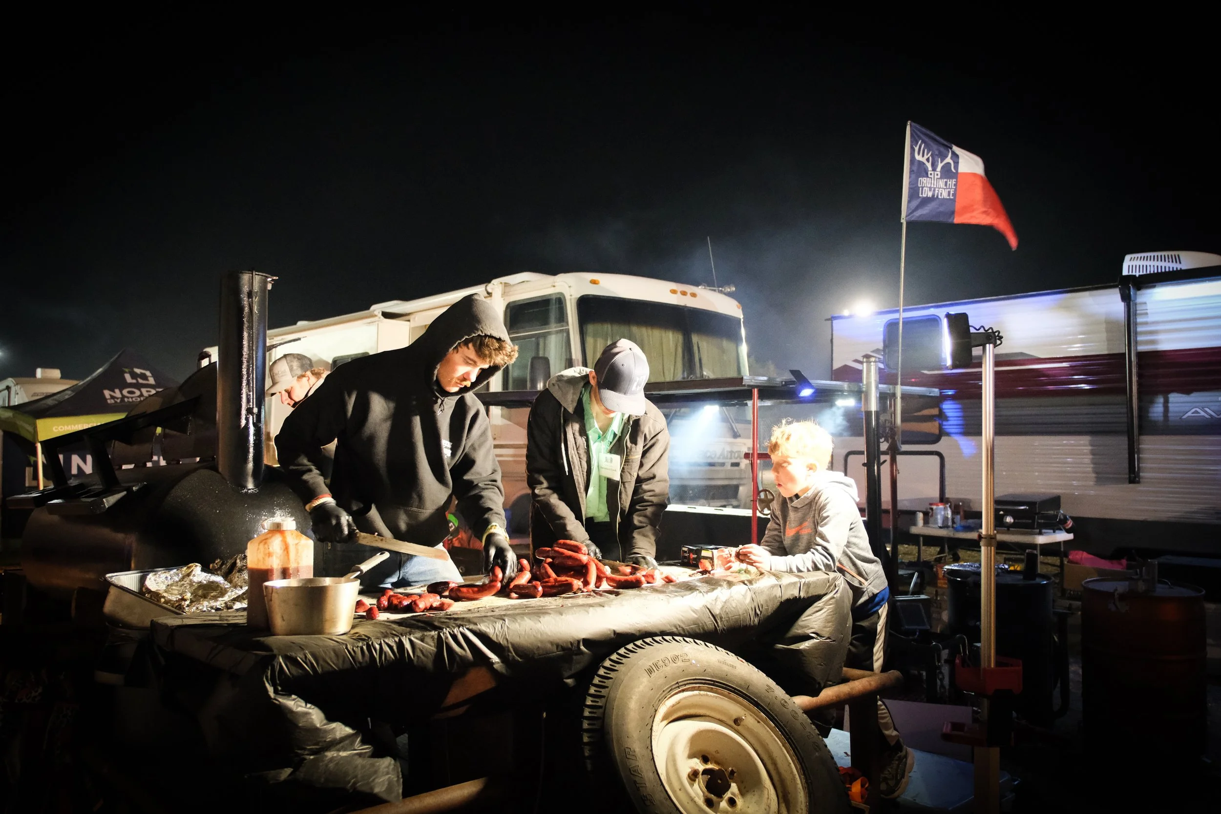 People grilling sausages at night in an outdoor setting with RVs, a flag, and a night sky.