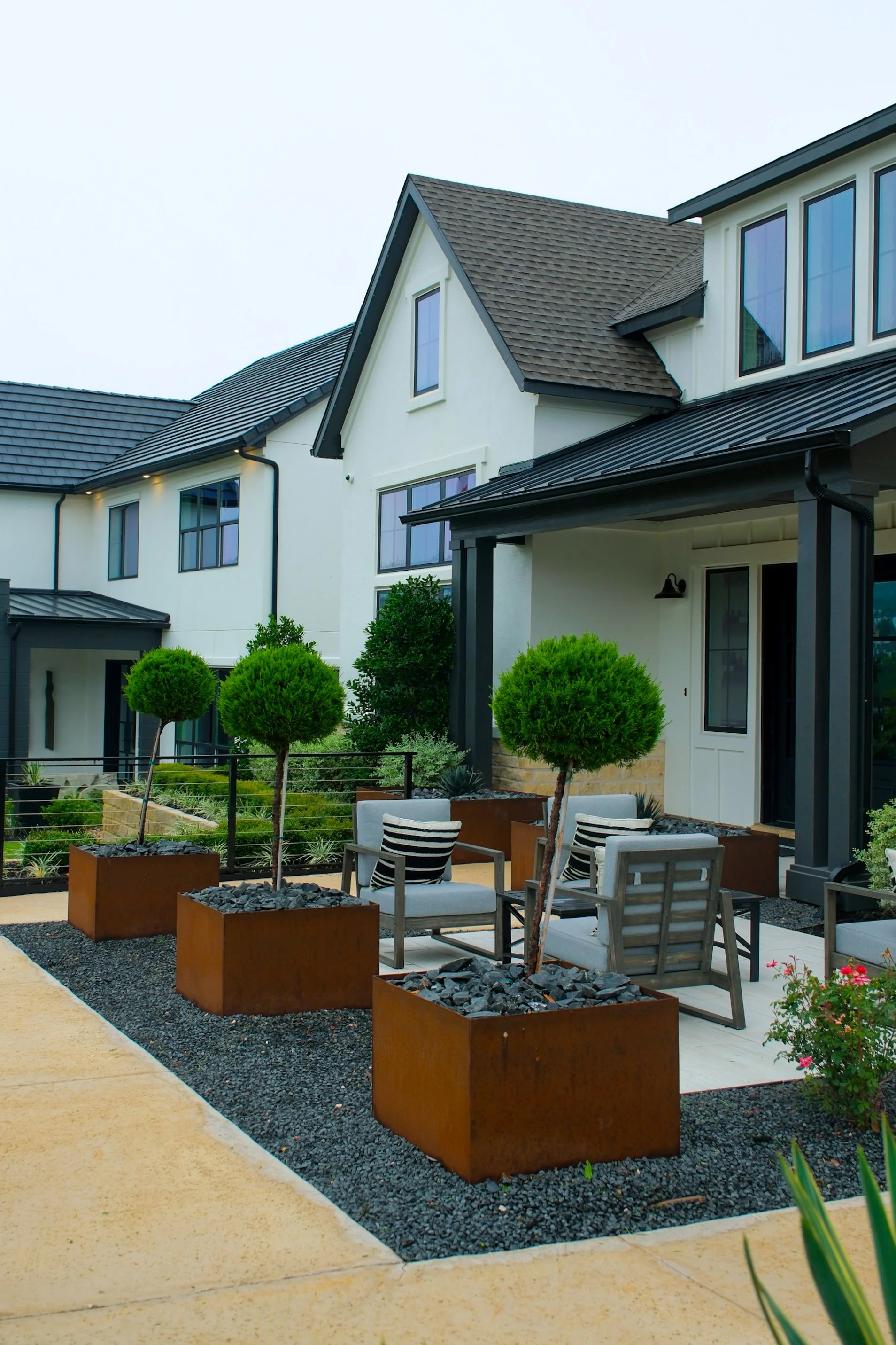 Modern house with a front porch, outdoor seating area with chairs, trees in rust-colored planters, and landscaped yard with plants and gravel.