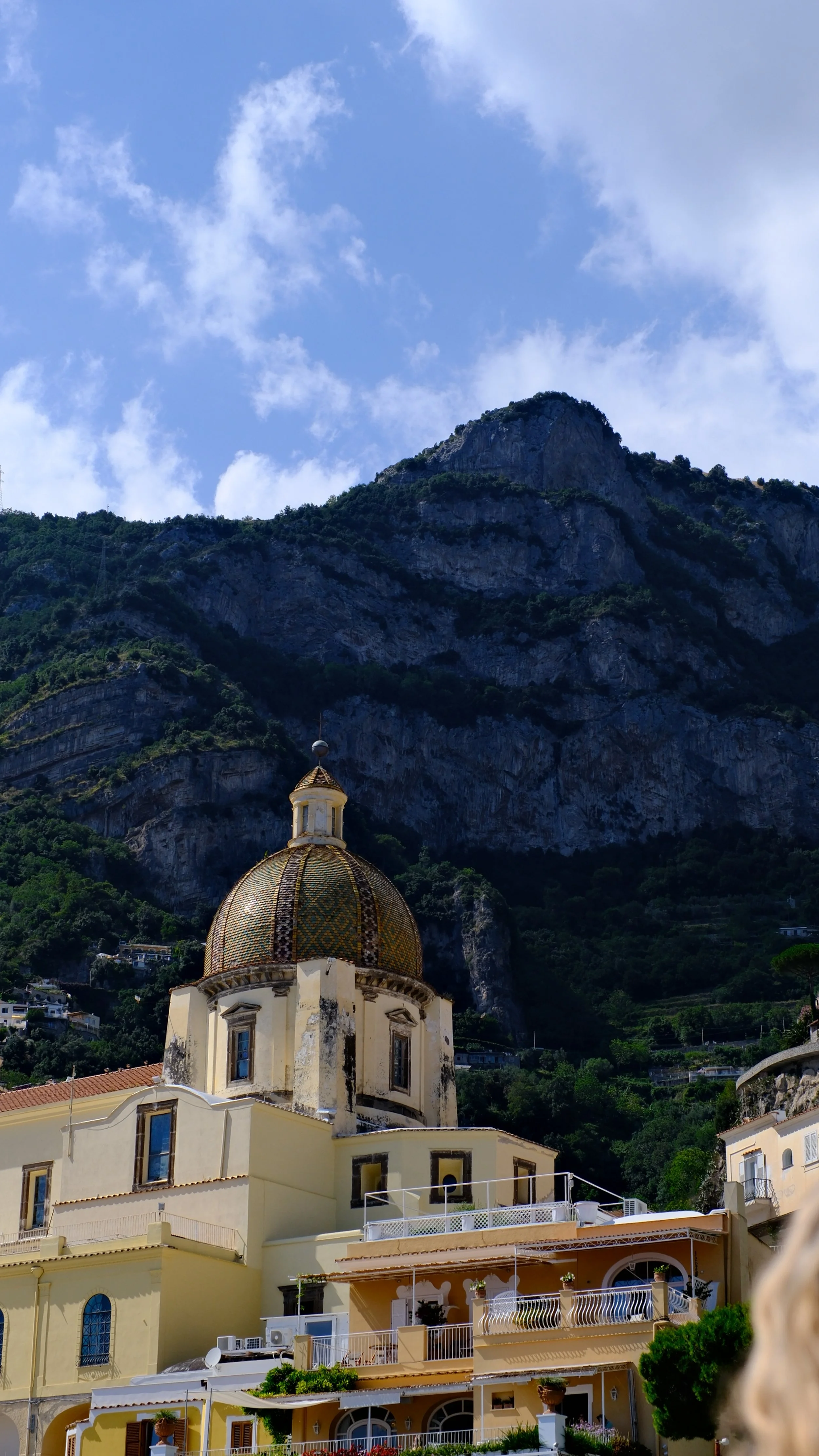 Colorful coastal village with a church featuring a domed roof, set against a steep hillside and rocky mountain with a partly cloudy sky.
