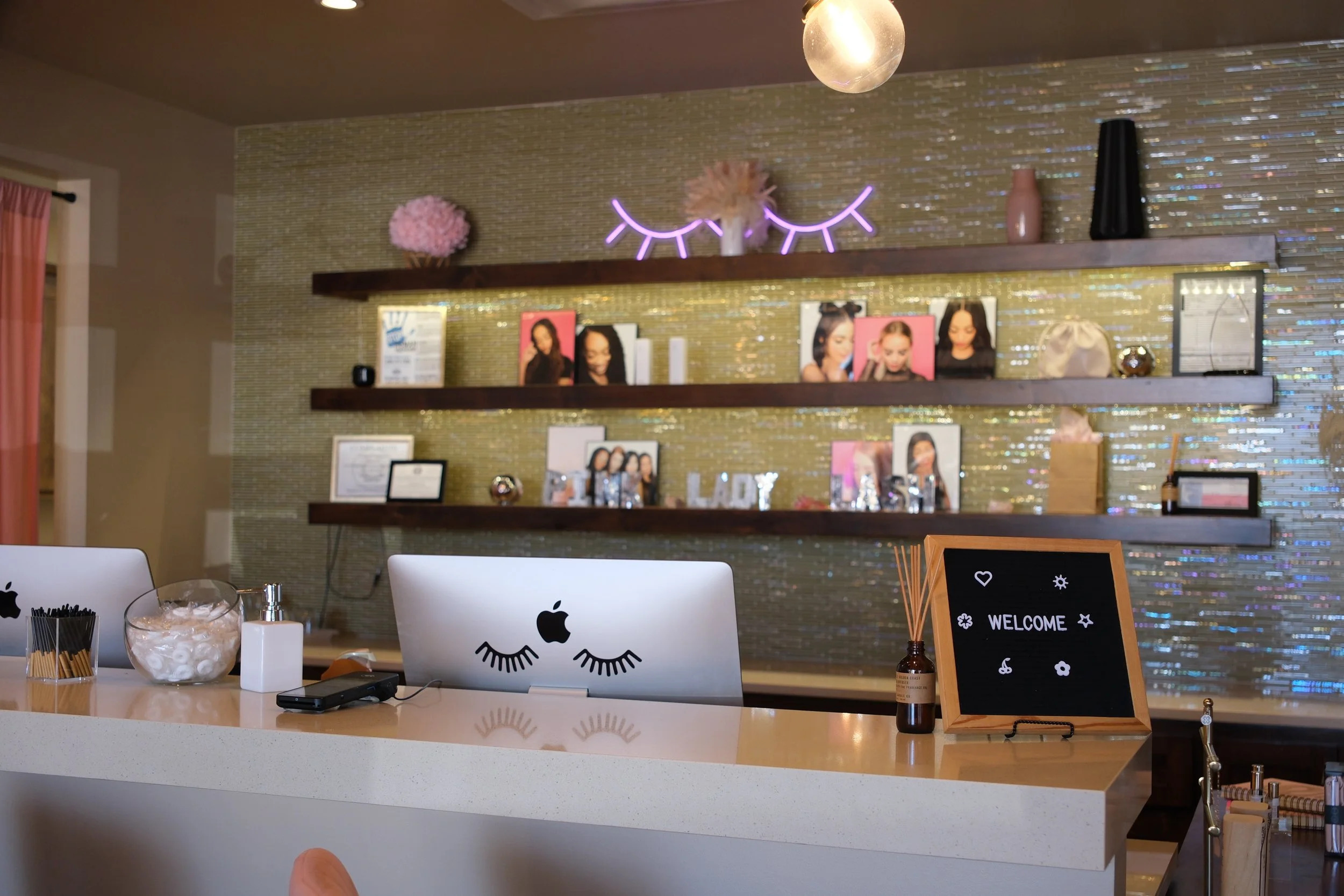 Reception desk with two Apple computers featuring eyelash decals, a bowl of cotton balls, a white bottle, and a welcome sign. Behind the desk is a wall with three dark wood shelves holding framed photos, vases, and decorative items. A selfie light an