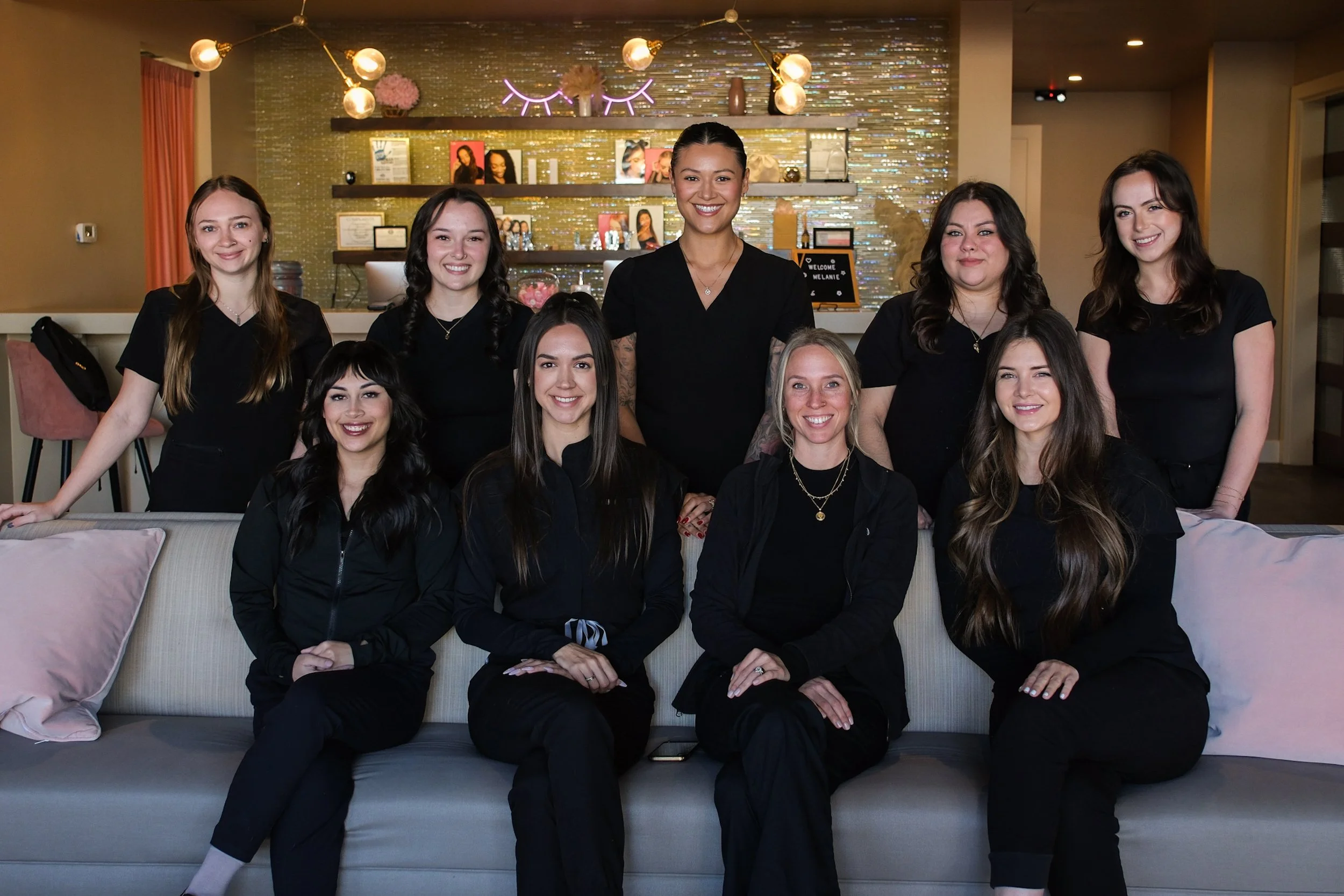 Group of nine women sitting and standing in a living room or salon with modern decor, smiling at the camera, all dressed in black.