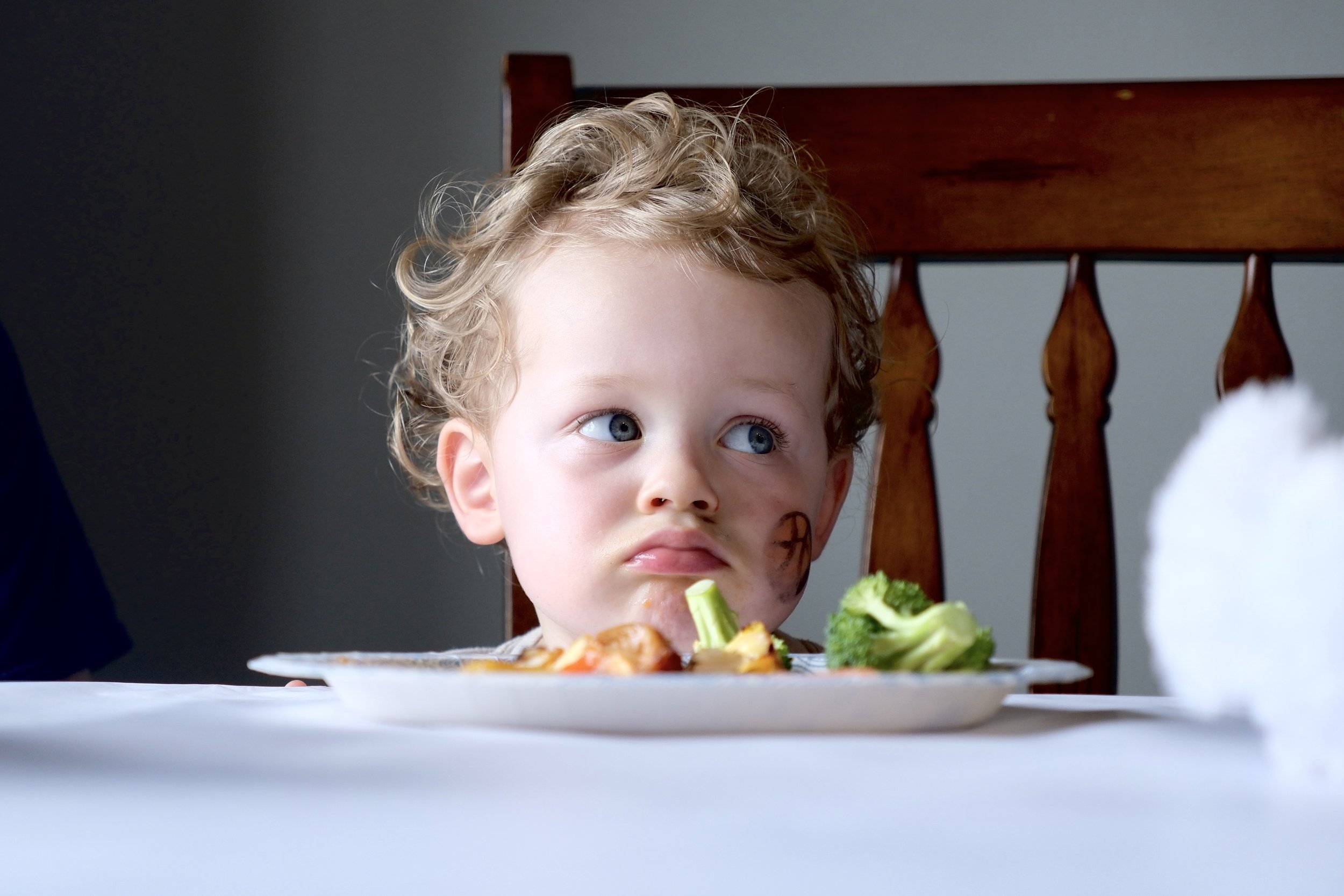 A young boy with curly blond hair sitting at a table with a plate of food, looking to the side with a pouty expression.