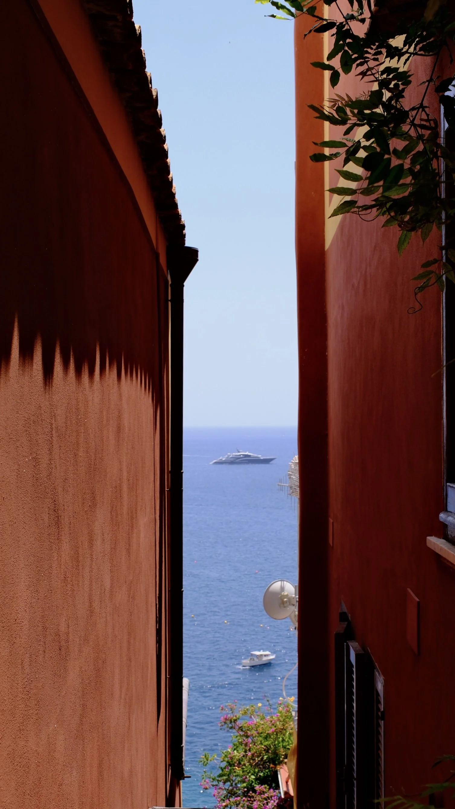 A narrow alley between two red stucco buildings with a view of the ocean, a large yacht, and smaller boats in the water.